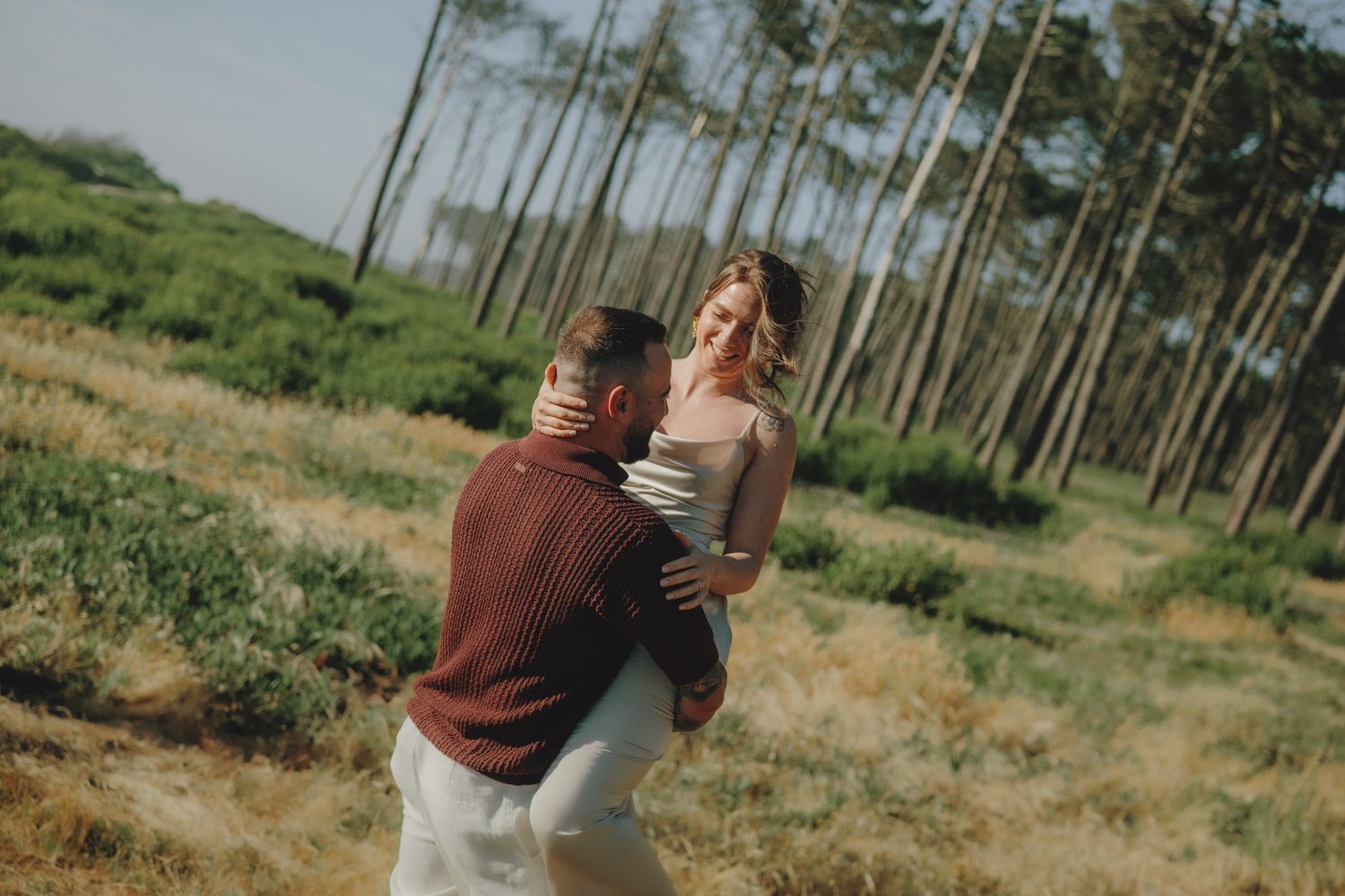 Playful couple embracing in pine forest during wedding photoshoot.