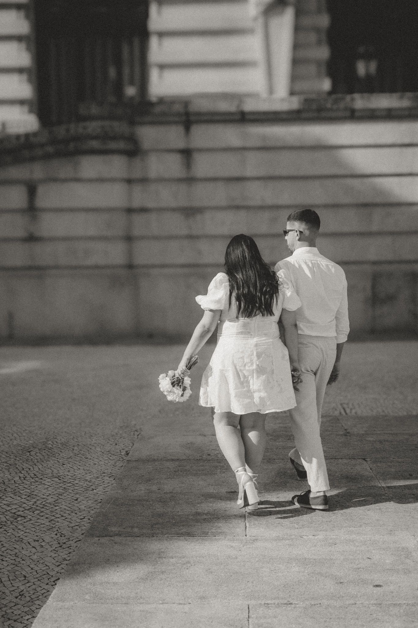 A couple in white outfits crouches together on a stone surface in a black and white photo.