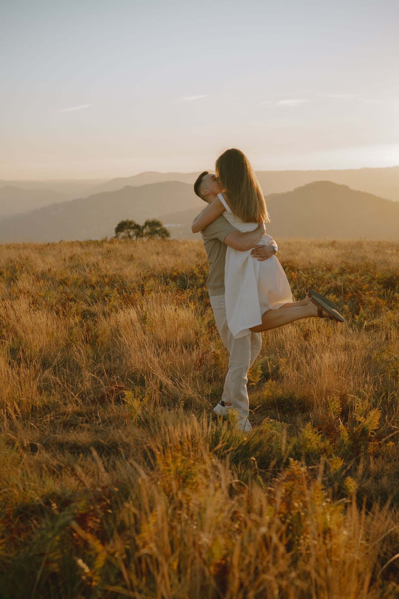 Two people share an intimate moment in a sun-drenched meadow with rolling hills behind them.