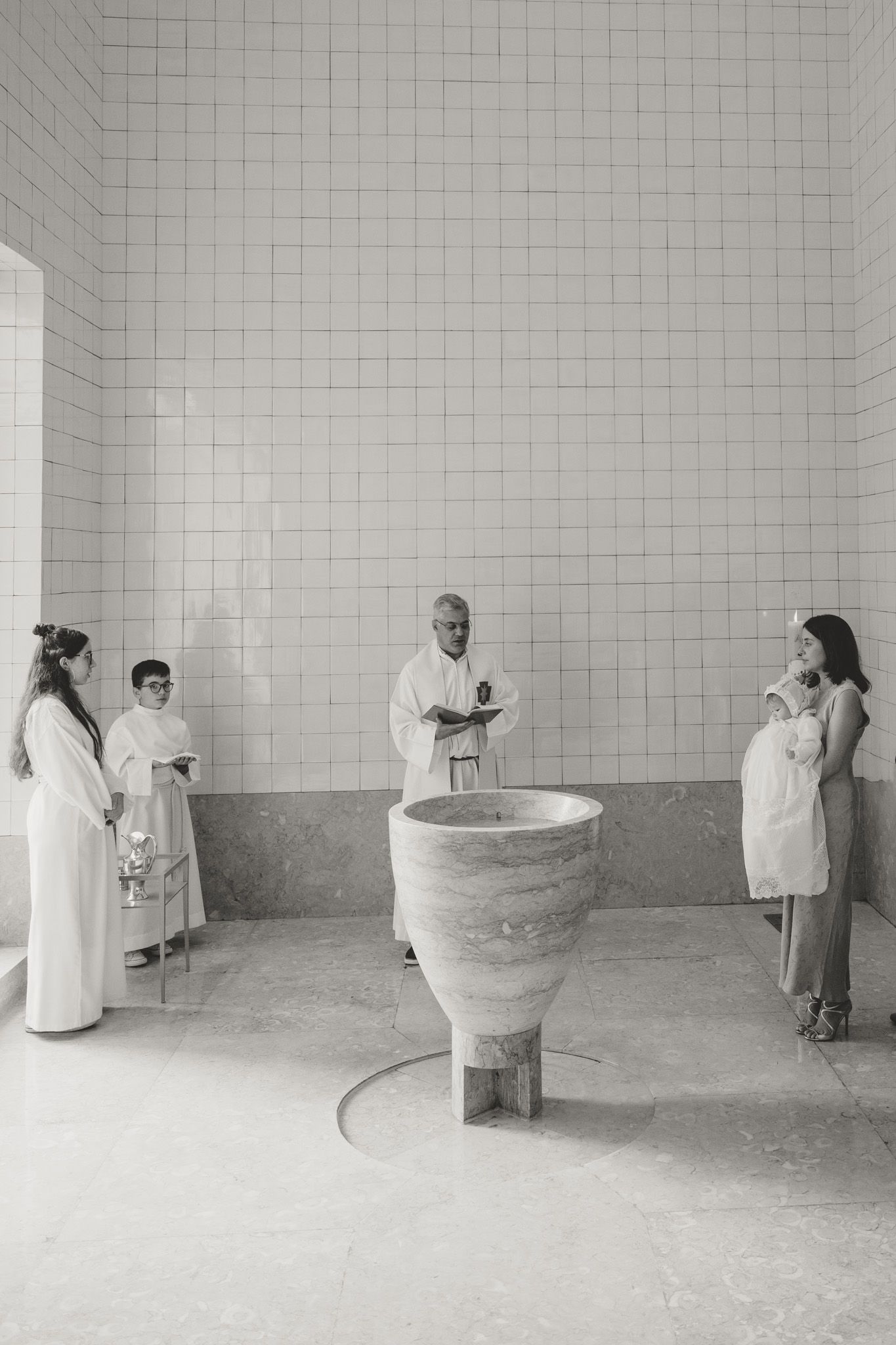 A sequence showing a baptismal ceremony in a minimalist white-tiled room with a stone font.