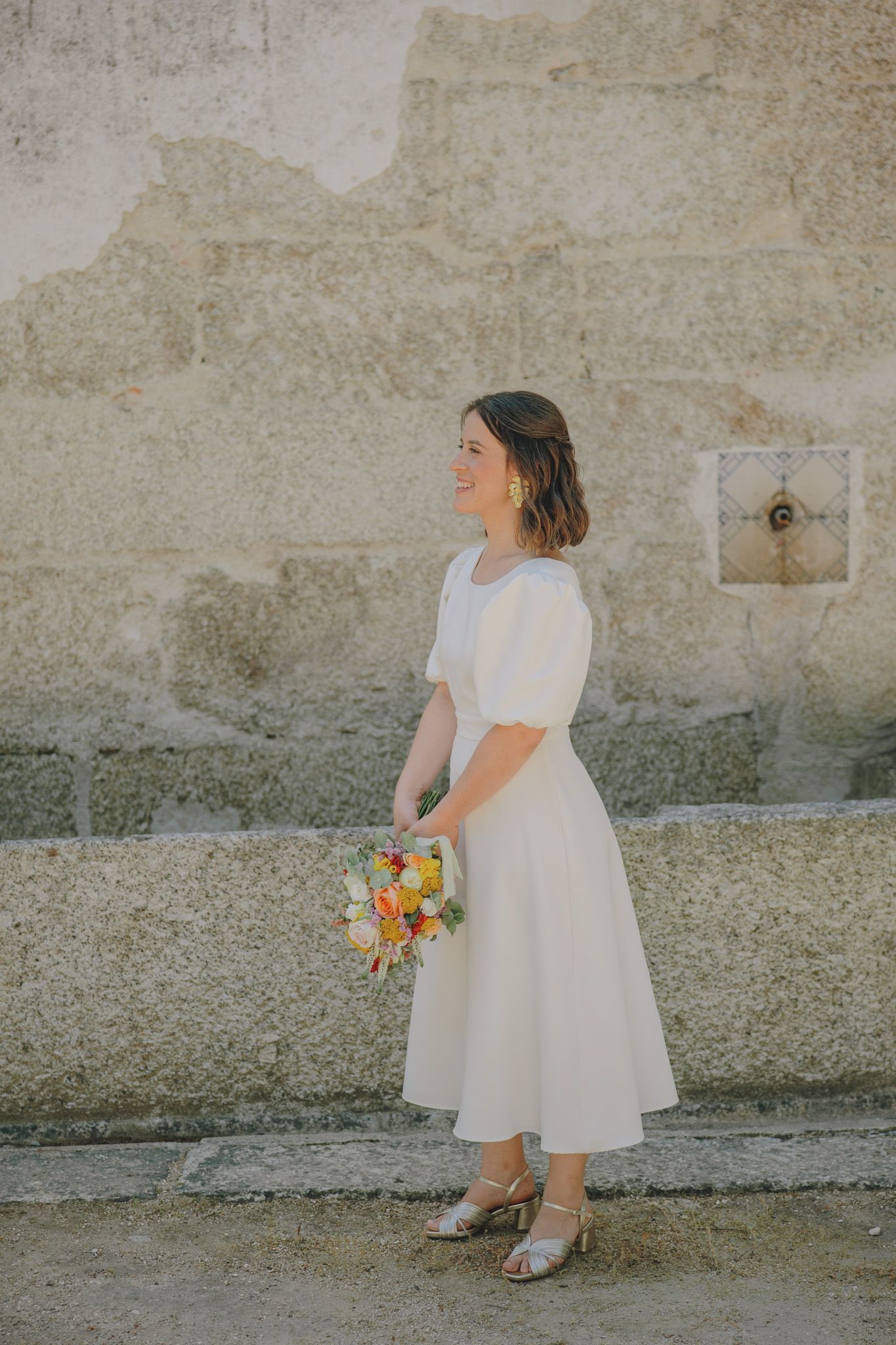 Young girl in white dress holding flowers against stone wall.