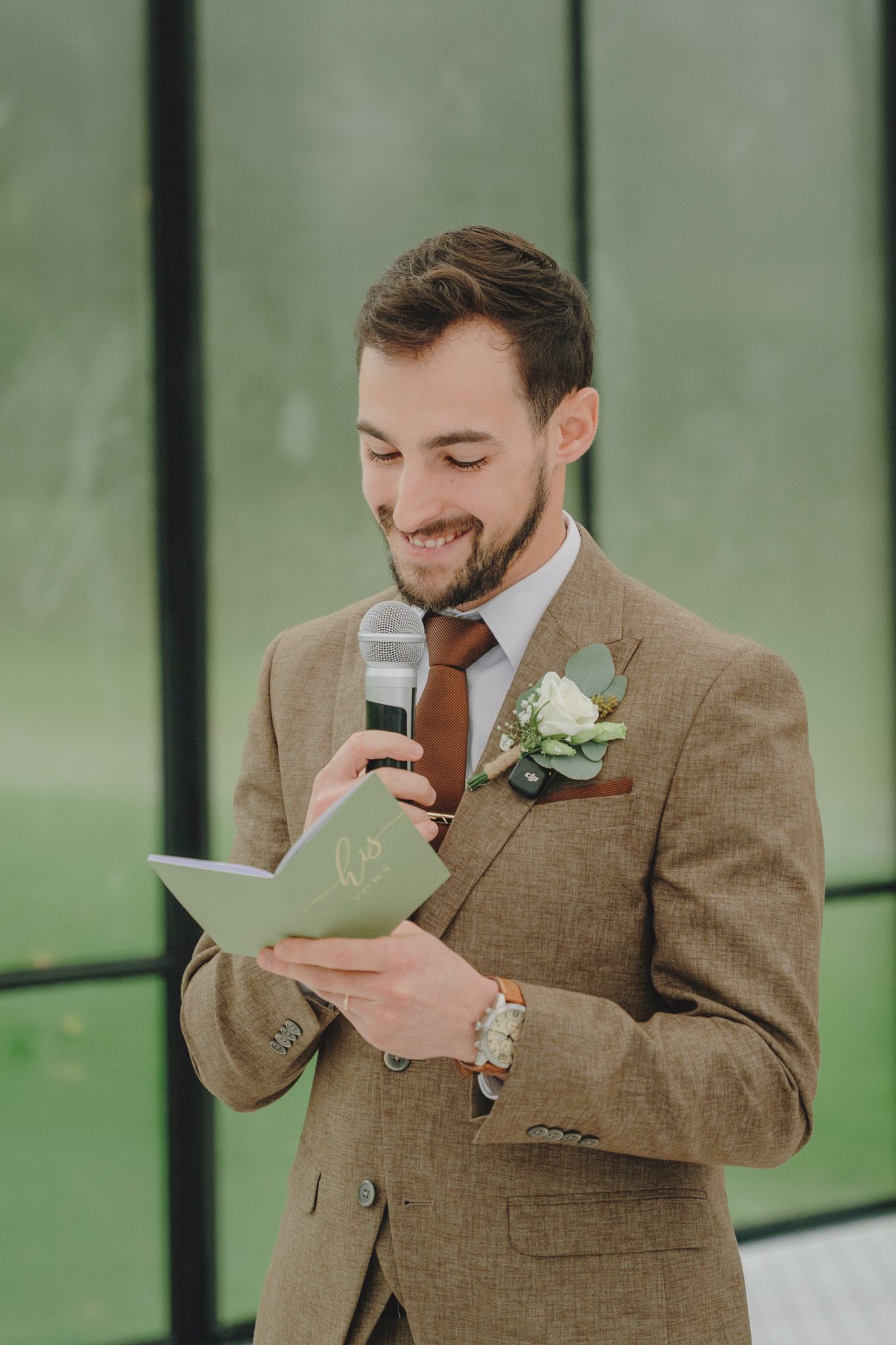 Groom in brown suit reads emotional vows from a card during wedding ceremony.