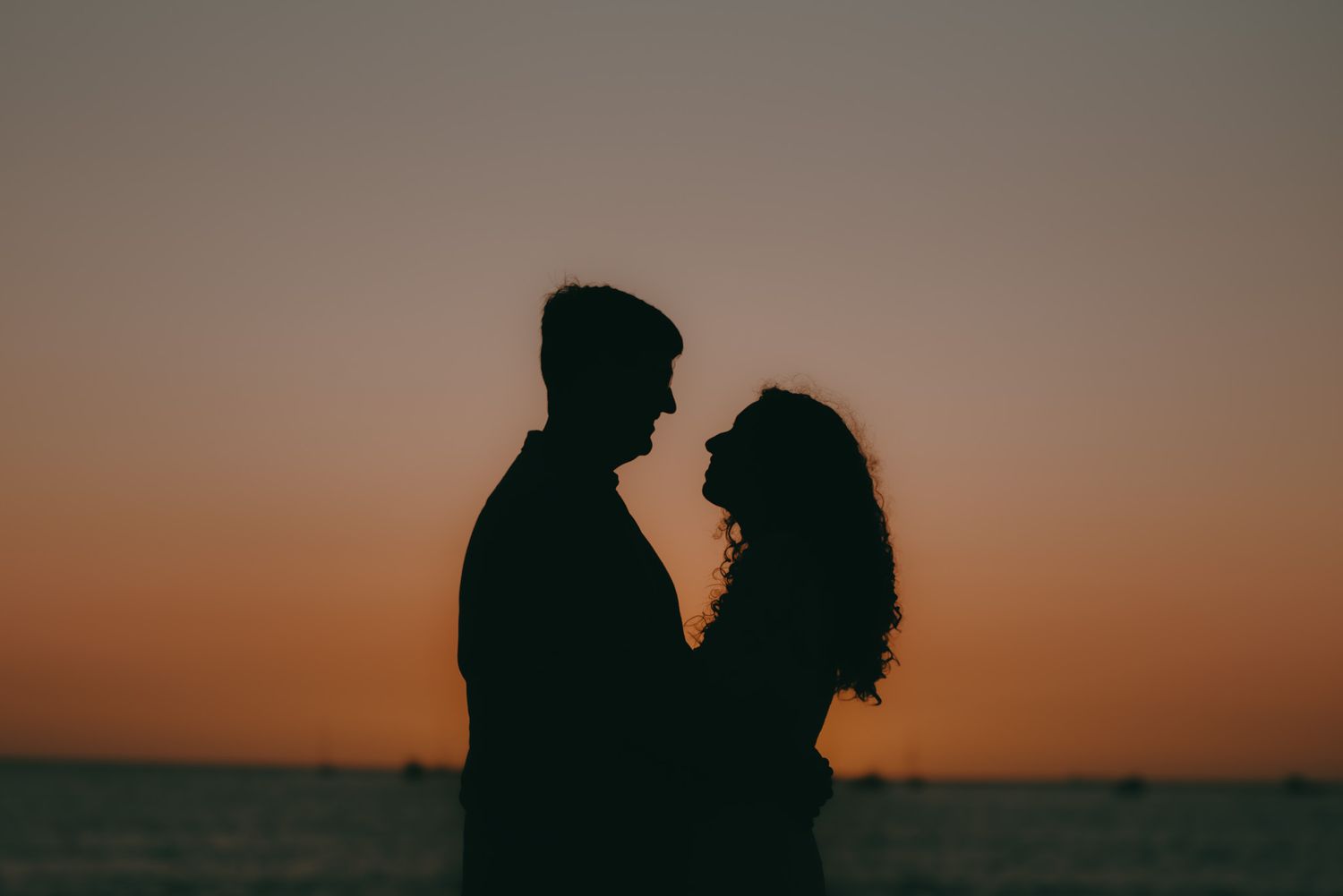 Dark silhouettes of a couple against an orange sunset sky over water.