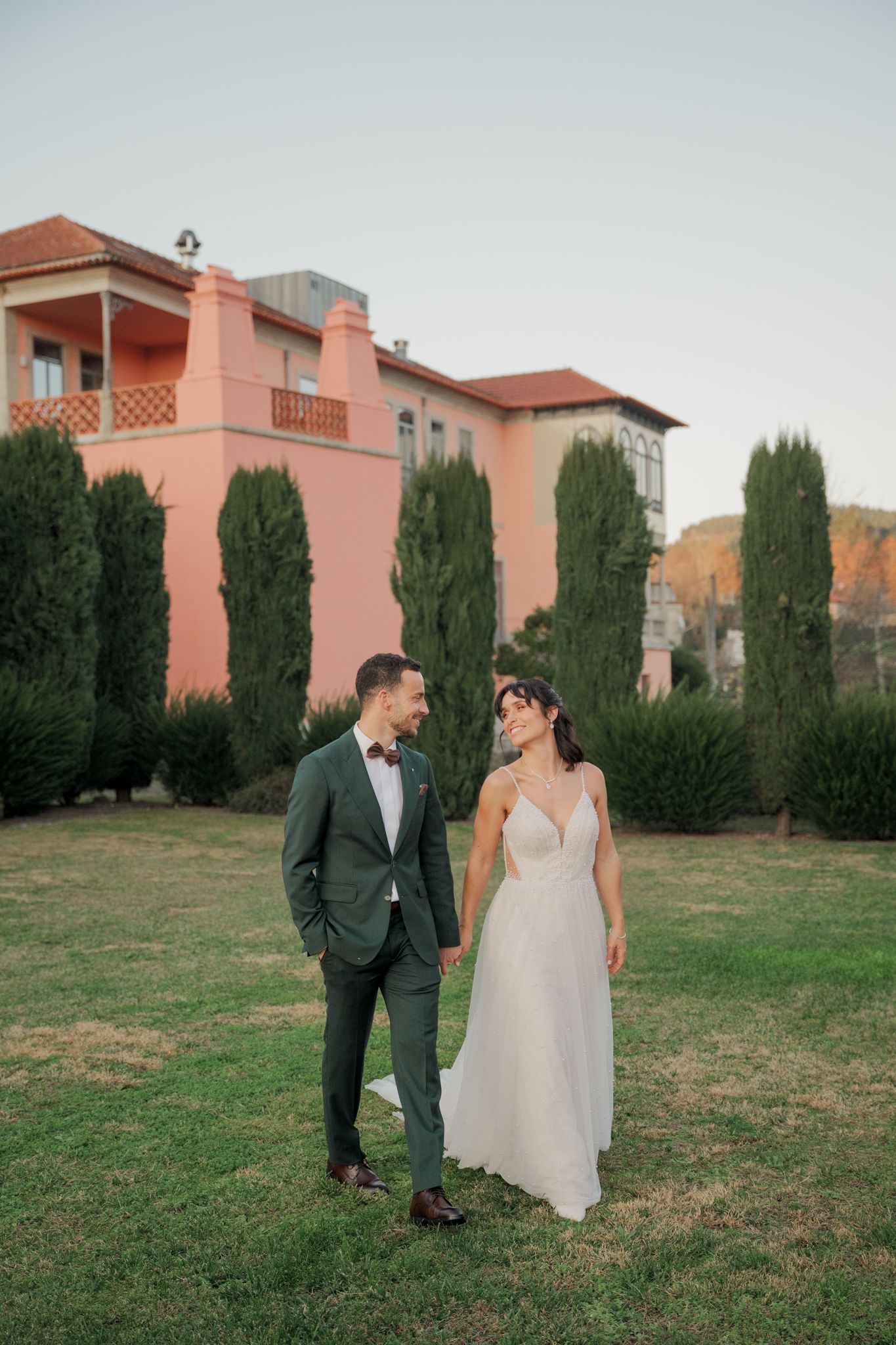 Newlyweds share intimate moment on lawn with pink Spanish-style architecture behind them.
