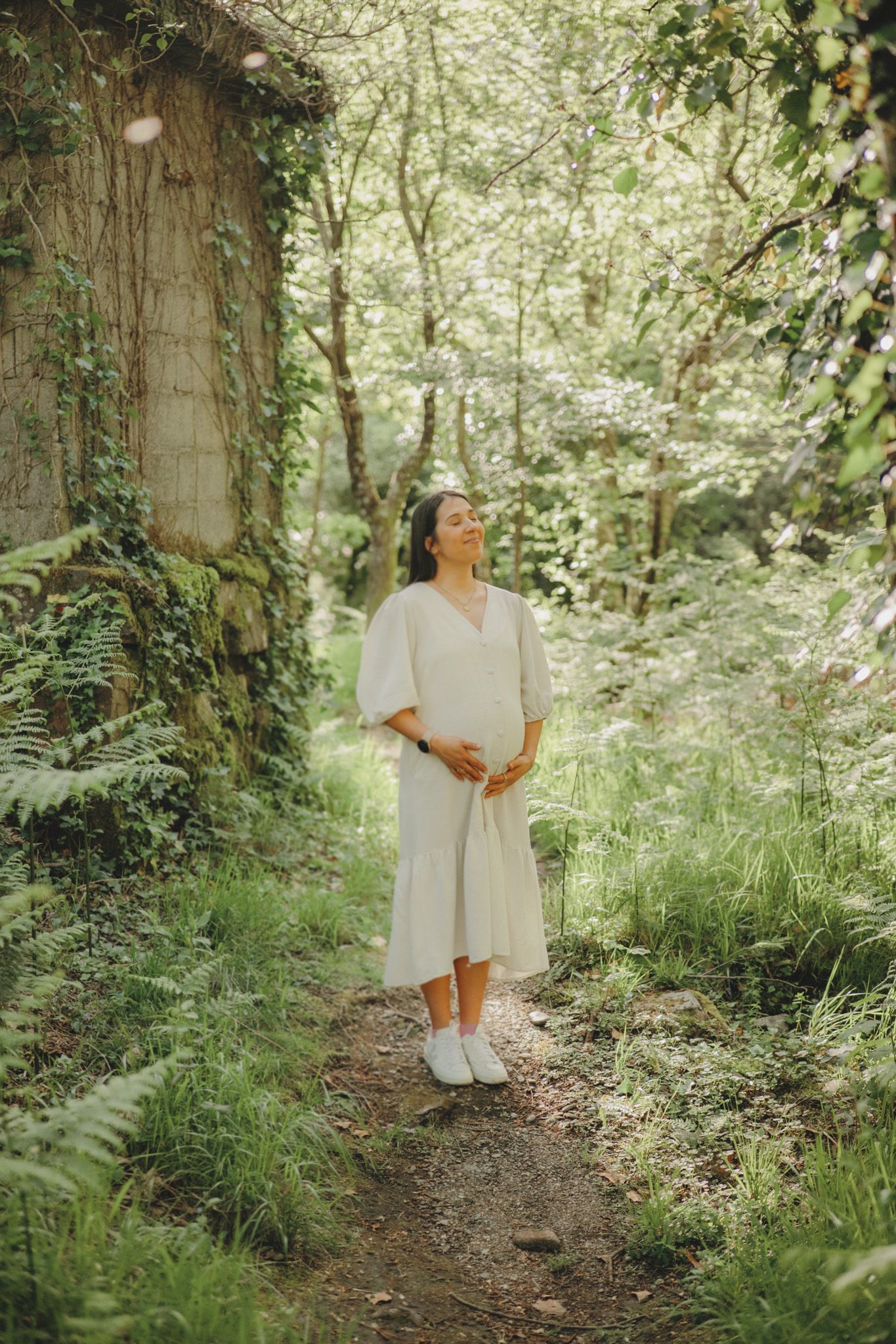 A person in white dress stands on a forest path surrounded by lush greenery and ivy-covered trees.