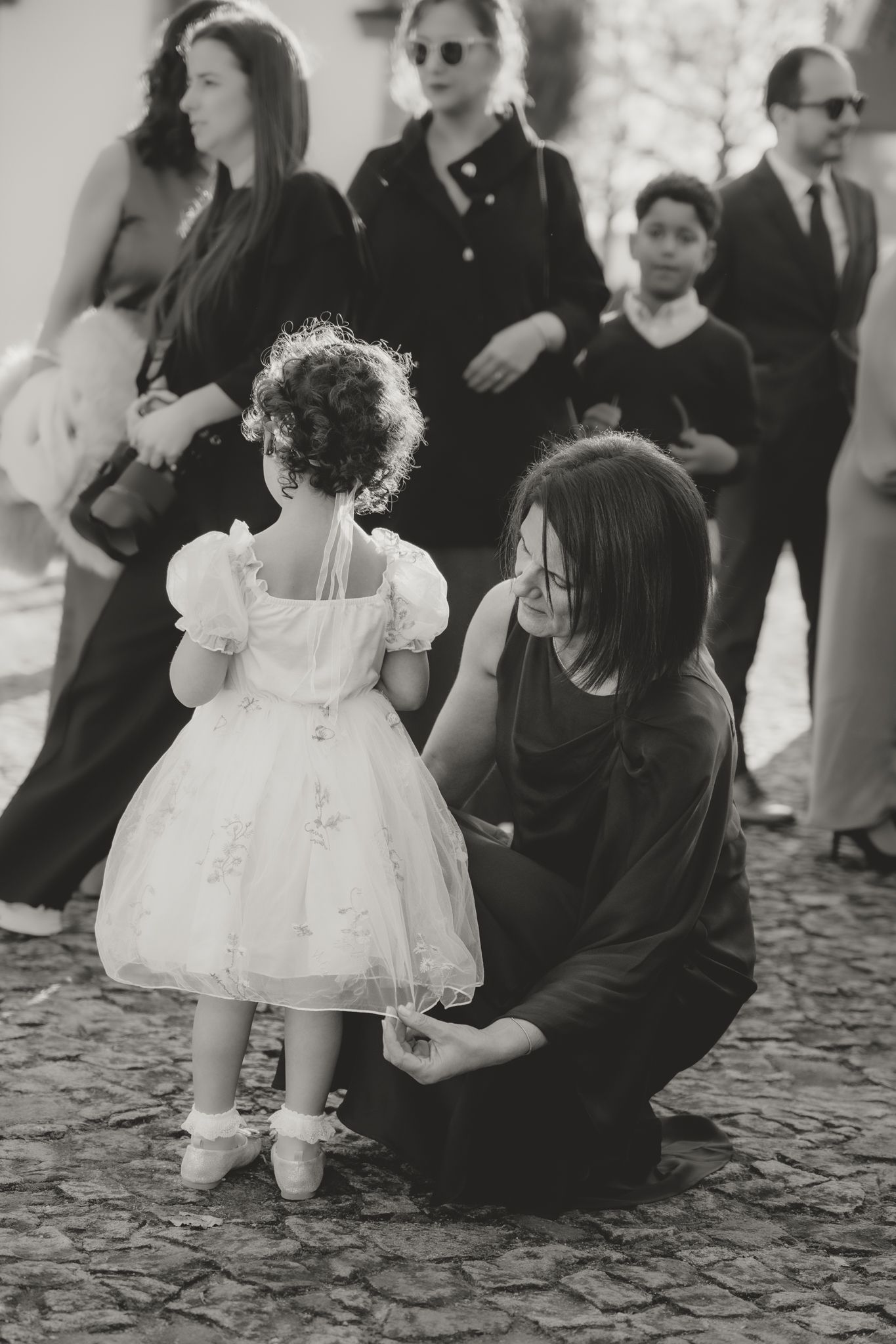 A small child in a white dress plays on cobblestone ground while adults watch during a formal event.