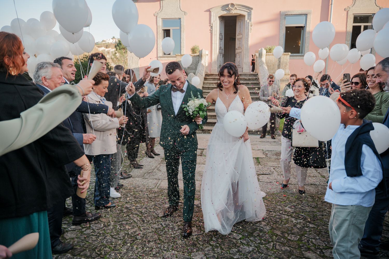 Joyful wedding party celebrating with white balloons outside vintage pink building.