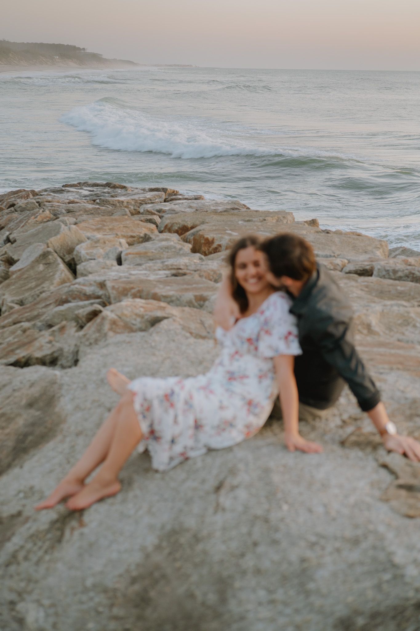 A person in a floral dress lies on coastal rocks at sunset while waves crash in the background.