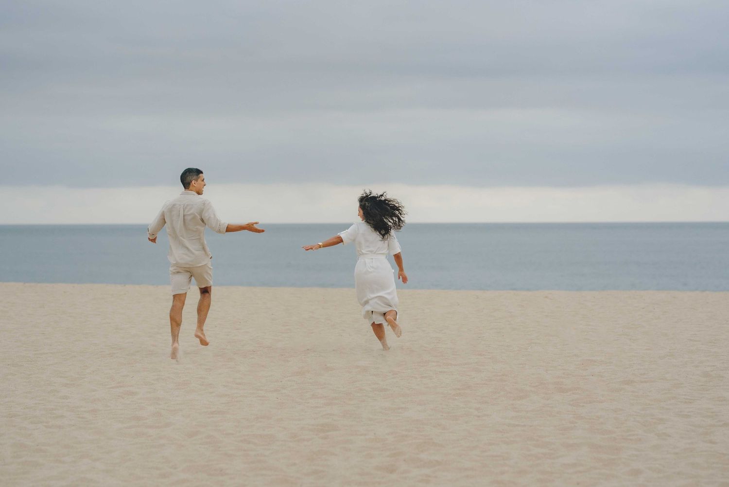 Two people joyfully leaping together on a sandy beach against an overcast ocean backdrop.