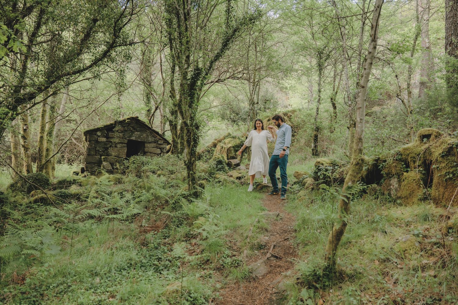 A couple walks along a forest path with a rustic wooden cabin in the background.