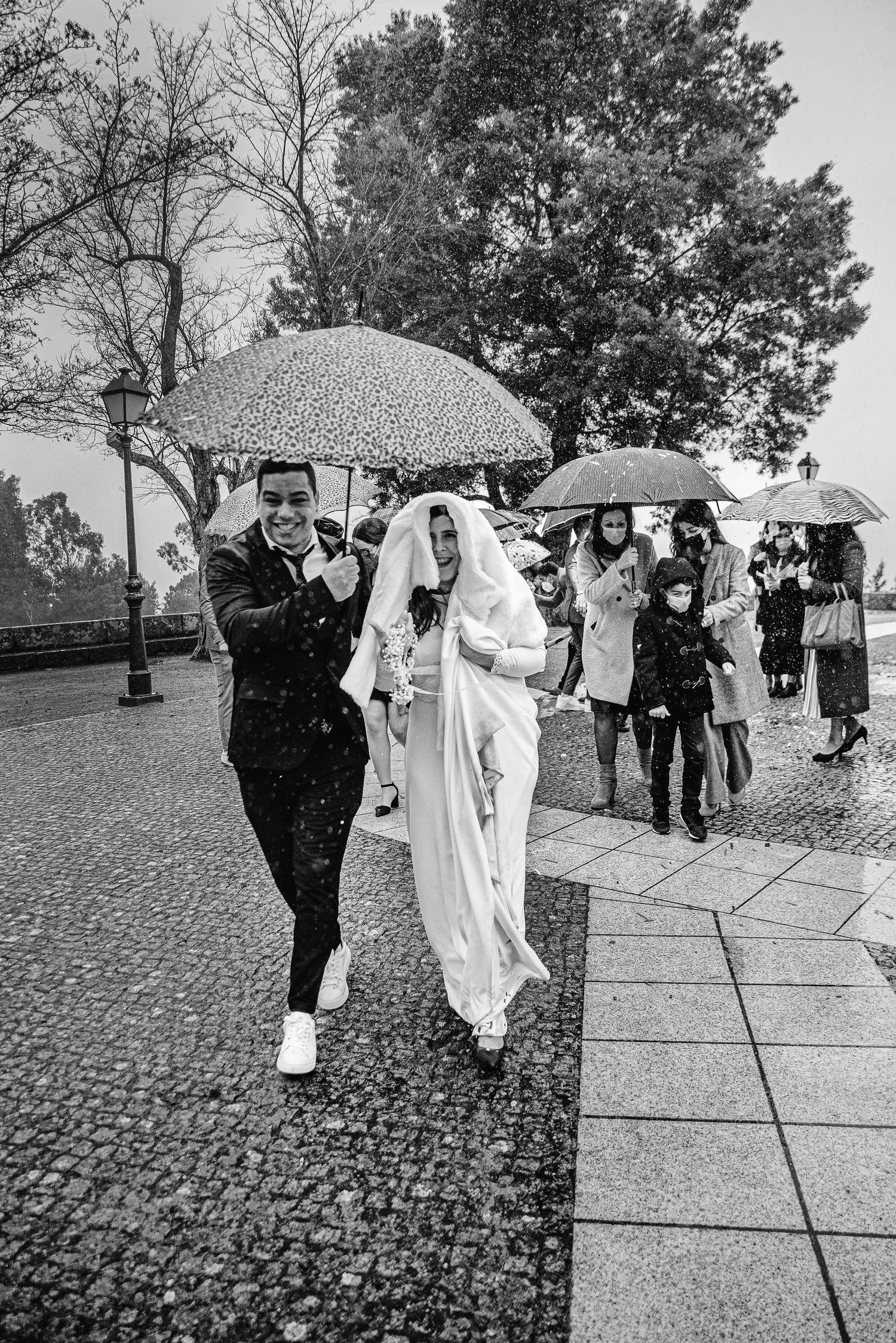 A wedding party walks under umbrellas on a rainy day along a cobblestone path.