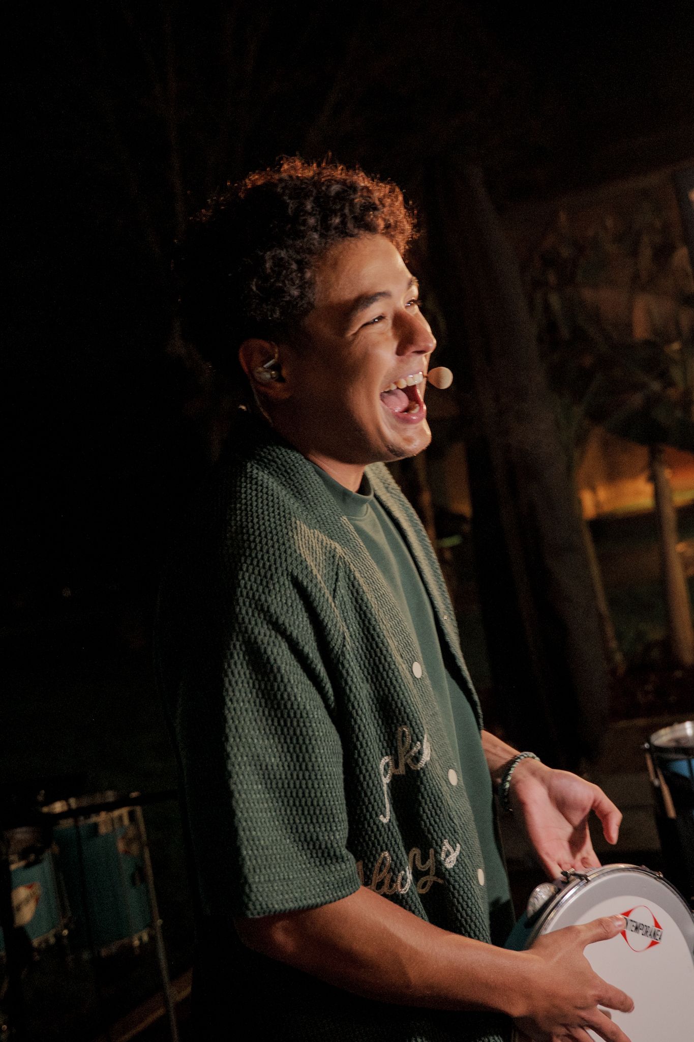 A person in green plays percussion instruments at a dimly lit event.