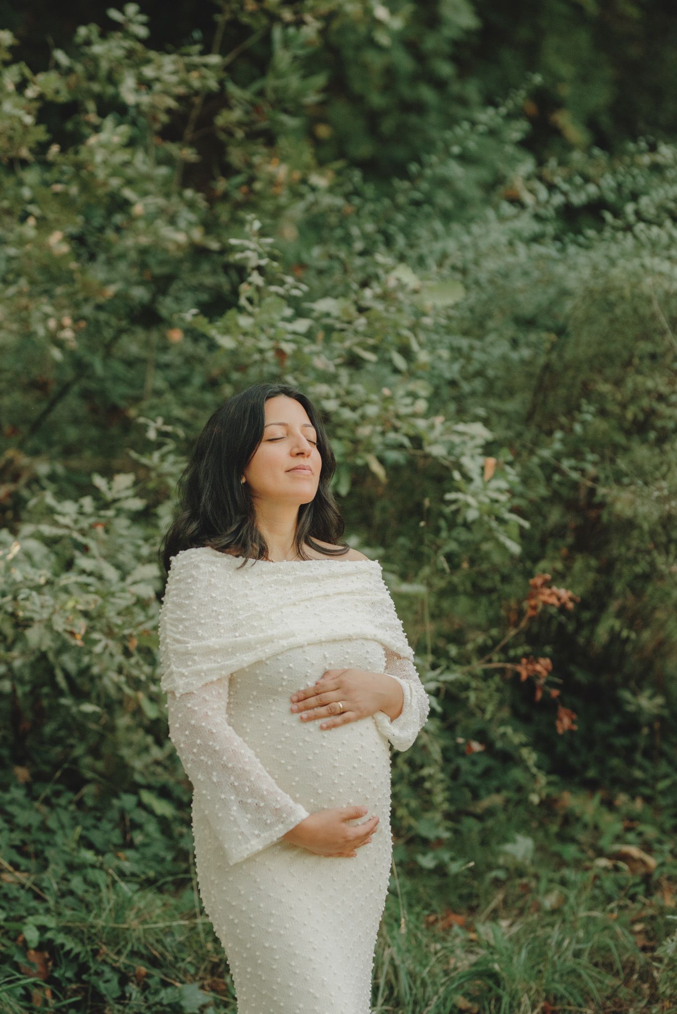Elegant maternity portrait in a white textured dress against lush green foliage.