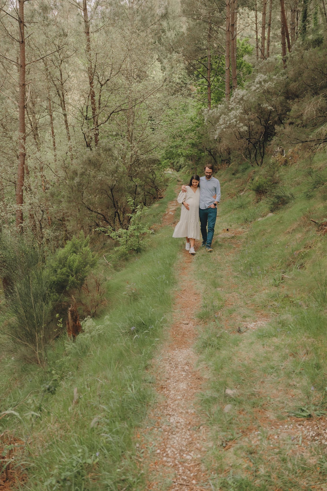Two people walking together on a winding forest trail surrounded by pine trees.