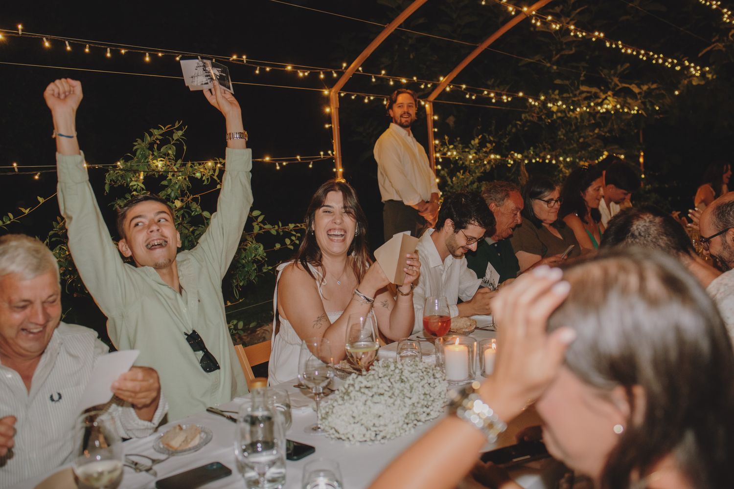 Wedding guests celebrating with raised glasses at an outdoor reception under string lights.