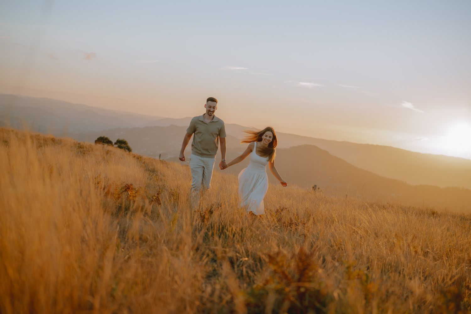 A couple walks hand in hand through a sun-soaked field with mountains in the distance.