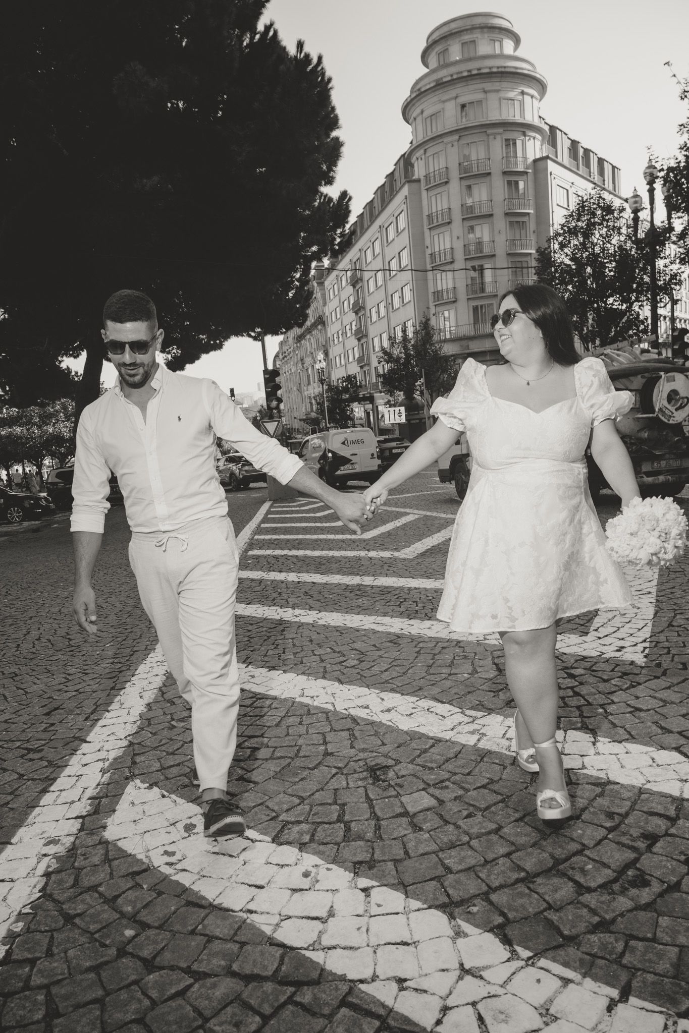 A couple in white clothing dances playfully on cobblestone streets.
