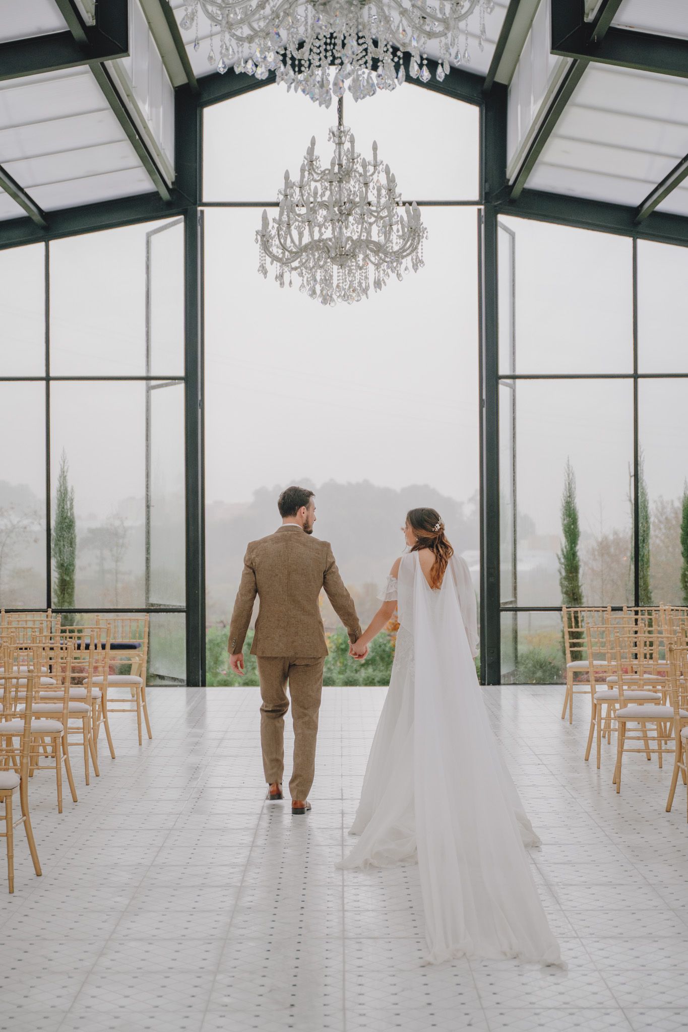 Elegant greenhouse wedding venue with crystal chandeliers and wooden chairs set for ceremony.