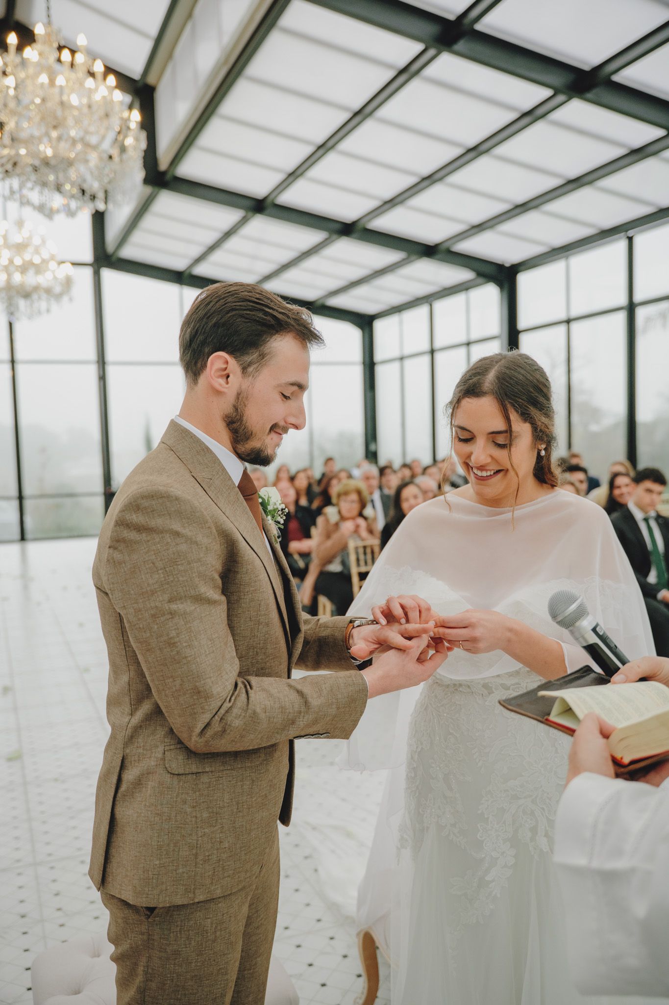 A couple exchanges rings during an intimate wedding ceremony in a sunlit greenhouse venue.