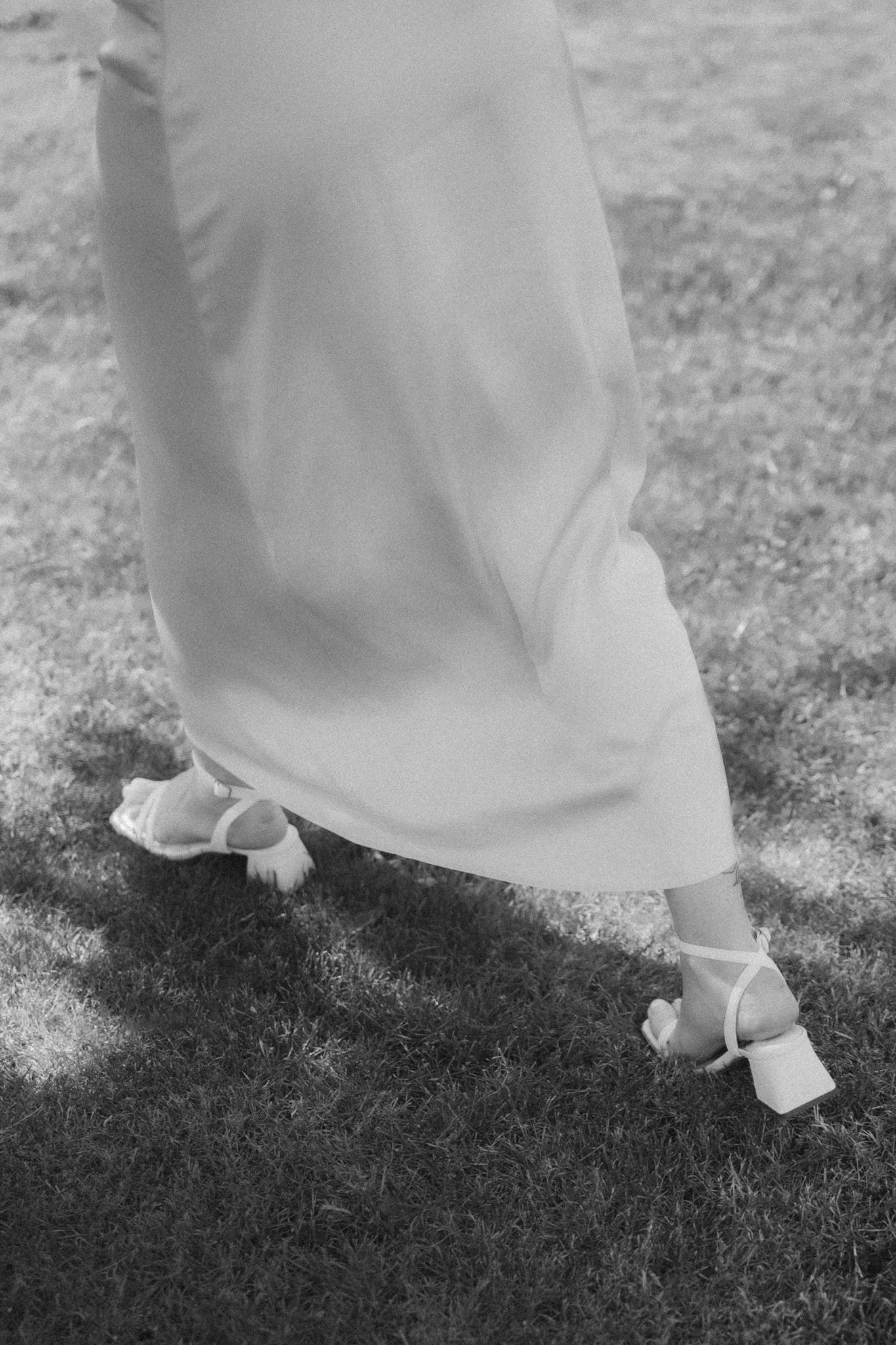 Close-up of elegant white bridal shoes and dress hem on grass in black and white.