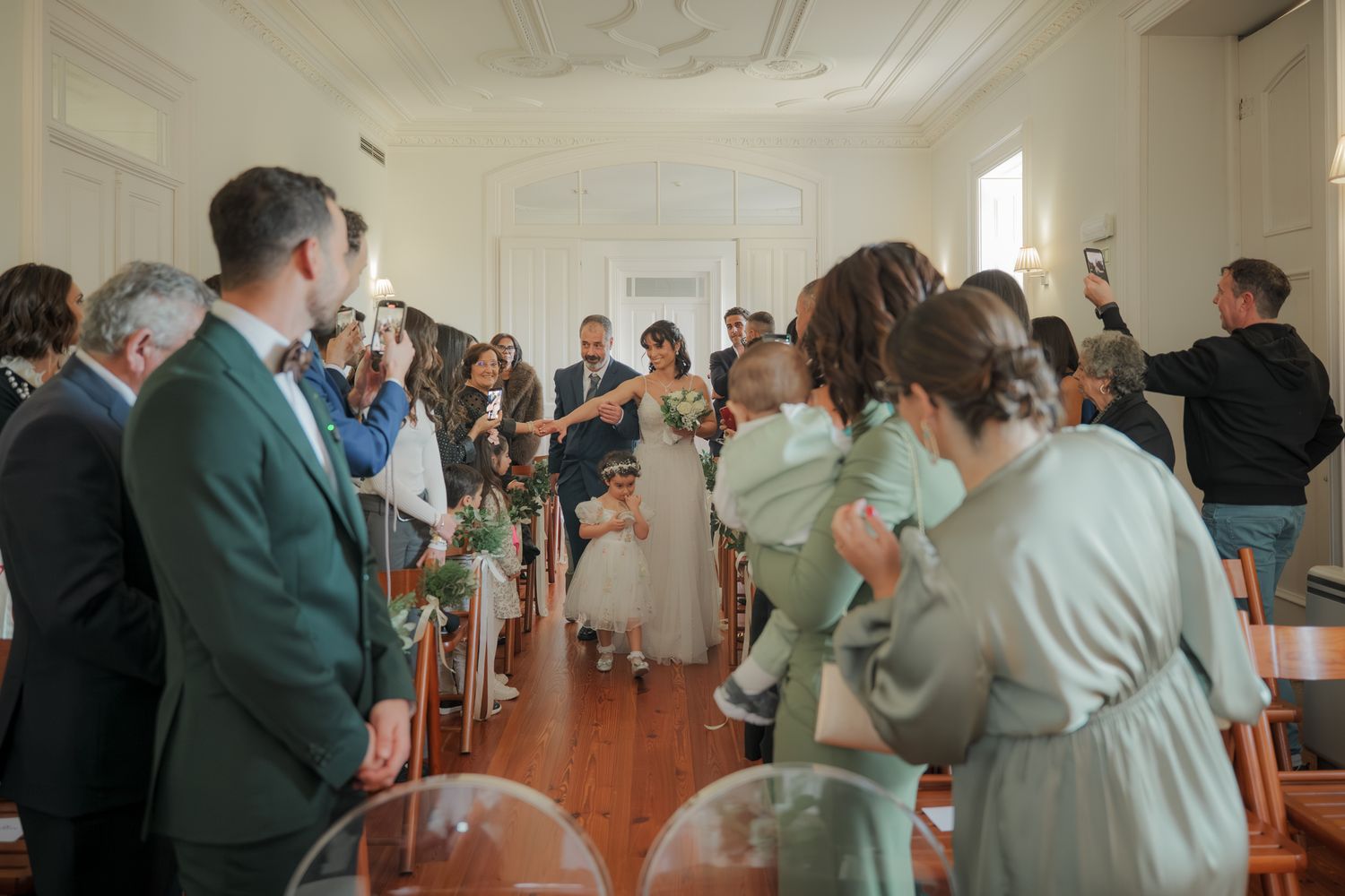 Wedding attendees stand in a bright indoor venue with decorative molding on the ceiling.