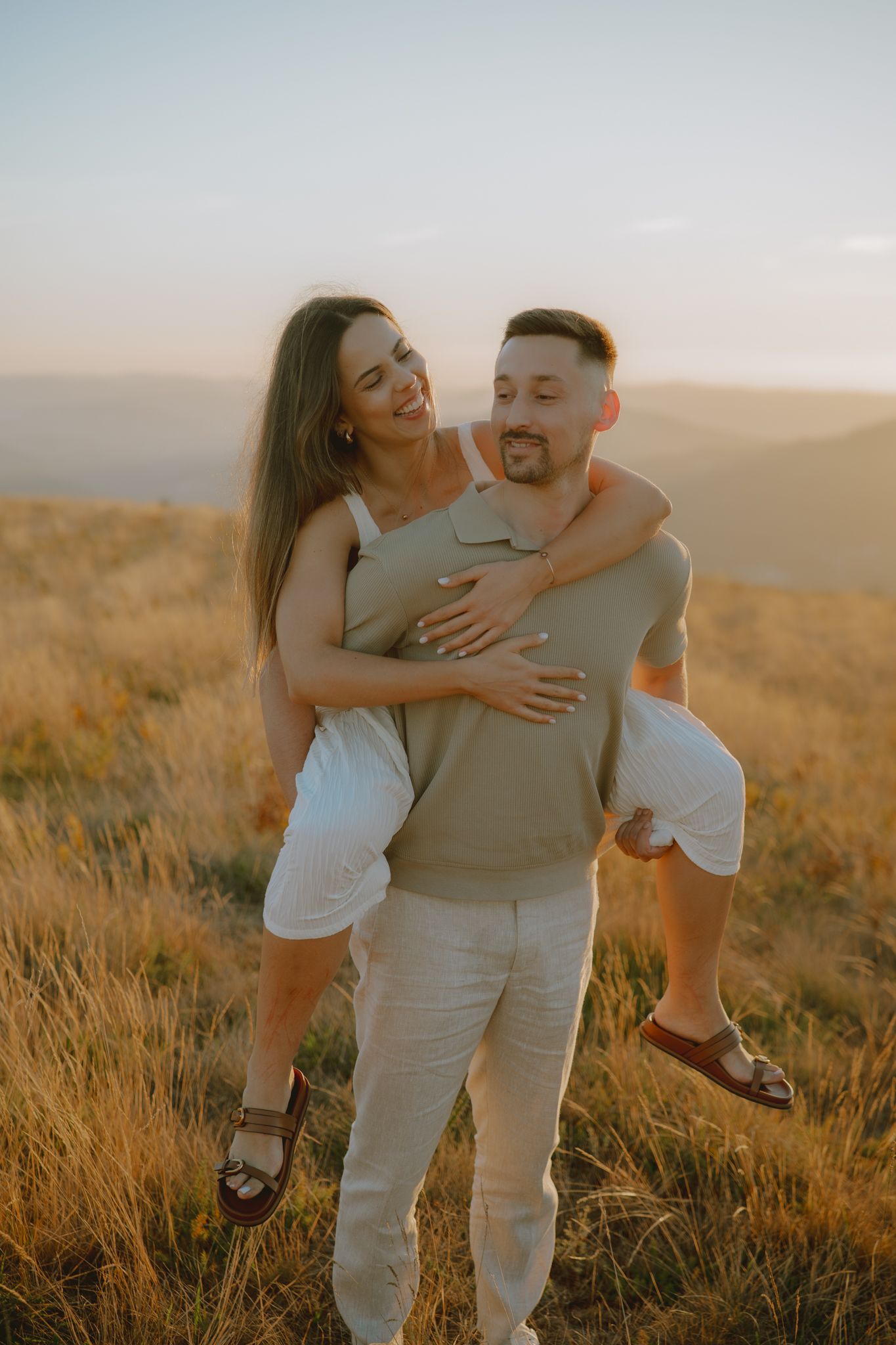 A playful couple shares laughter and embraces in tall grass during golden hour.