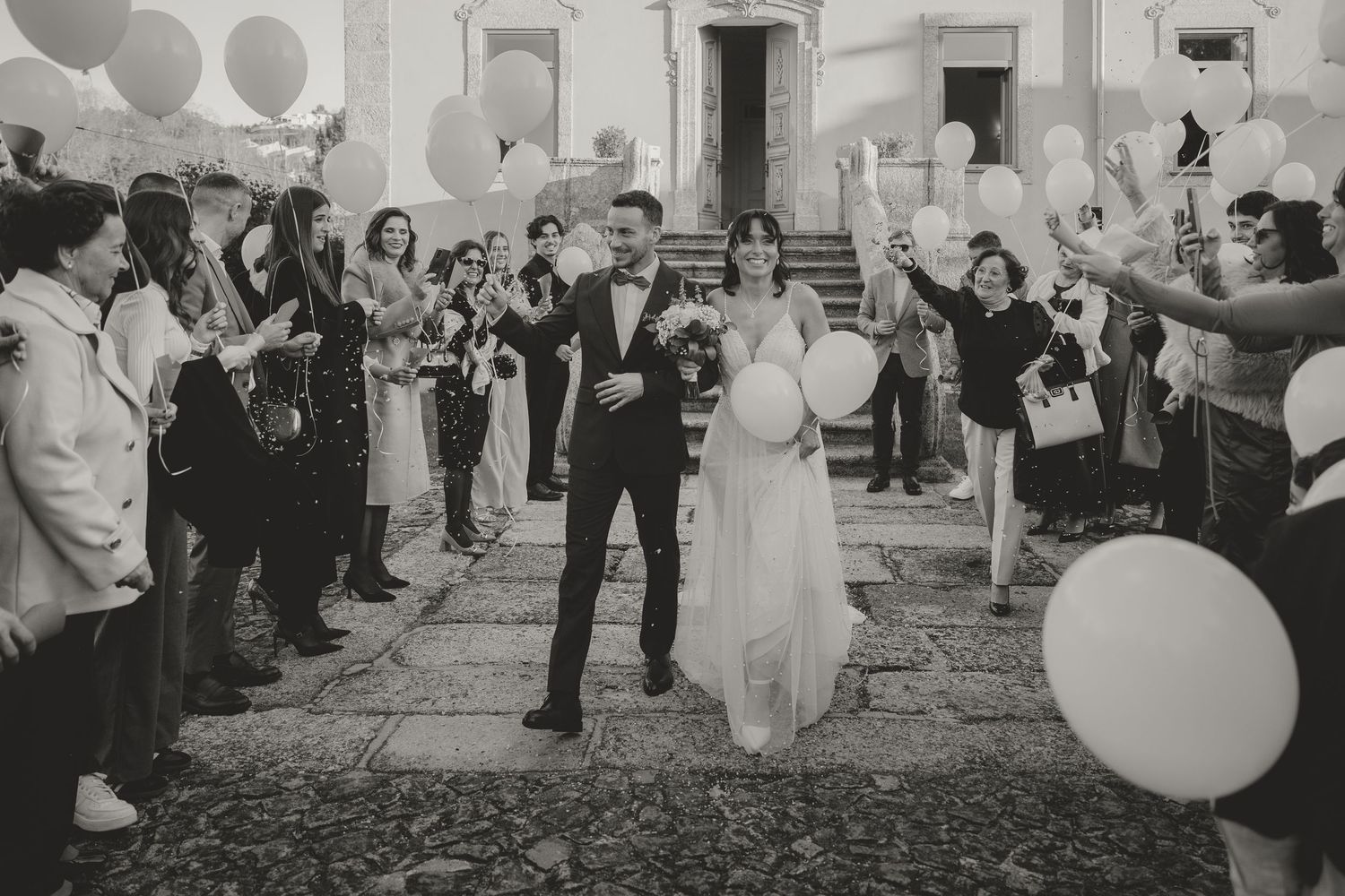 Wedding guests in formal attire hold white balloons while celebrating on a garden path during a festive reception.