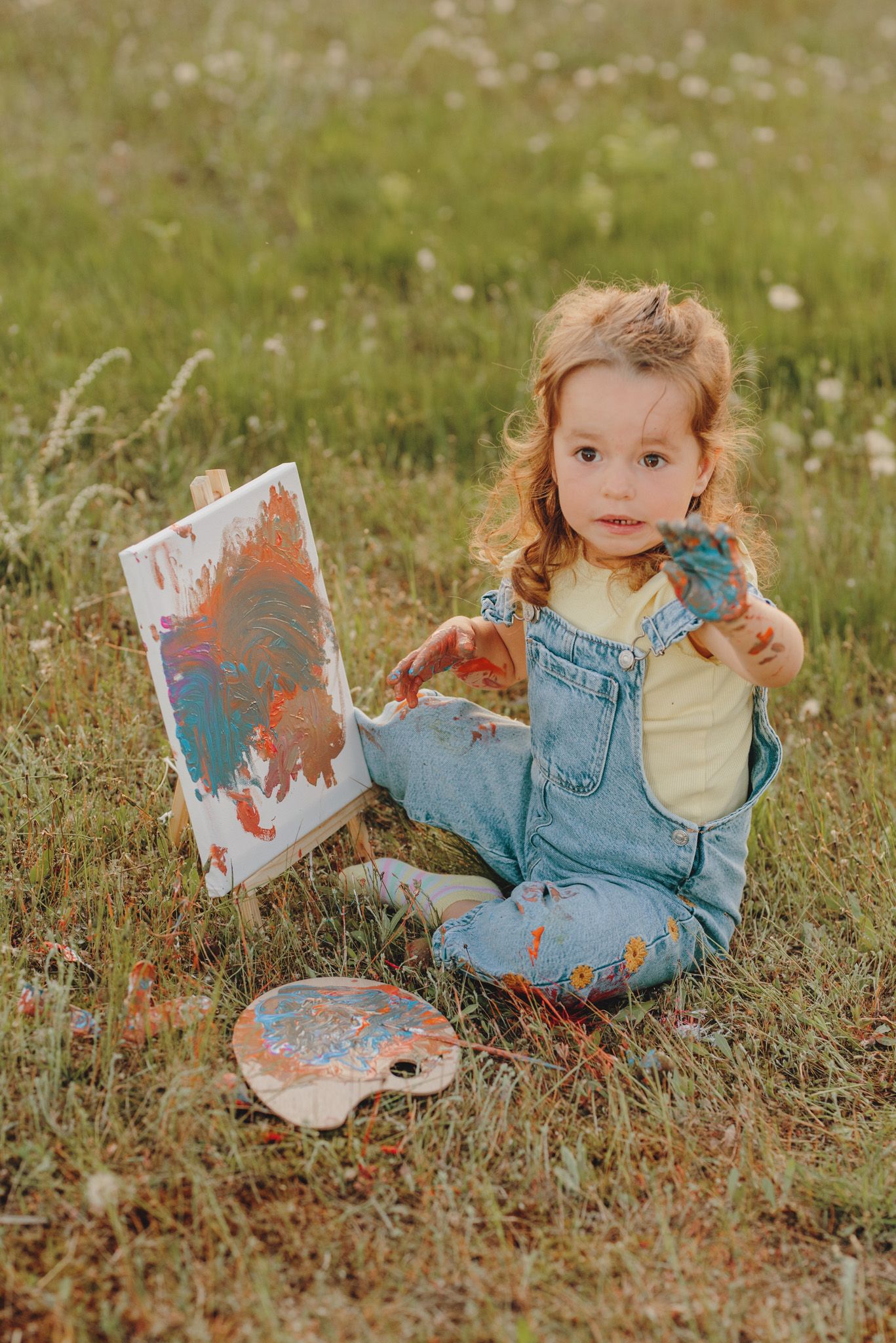 Small artist in denim overalls creates colorful artwork while sitting among wildflowers.