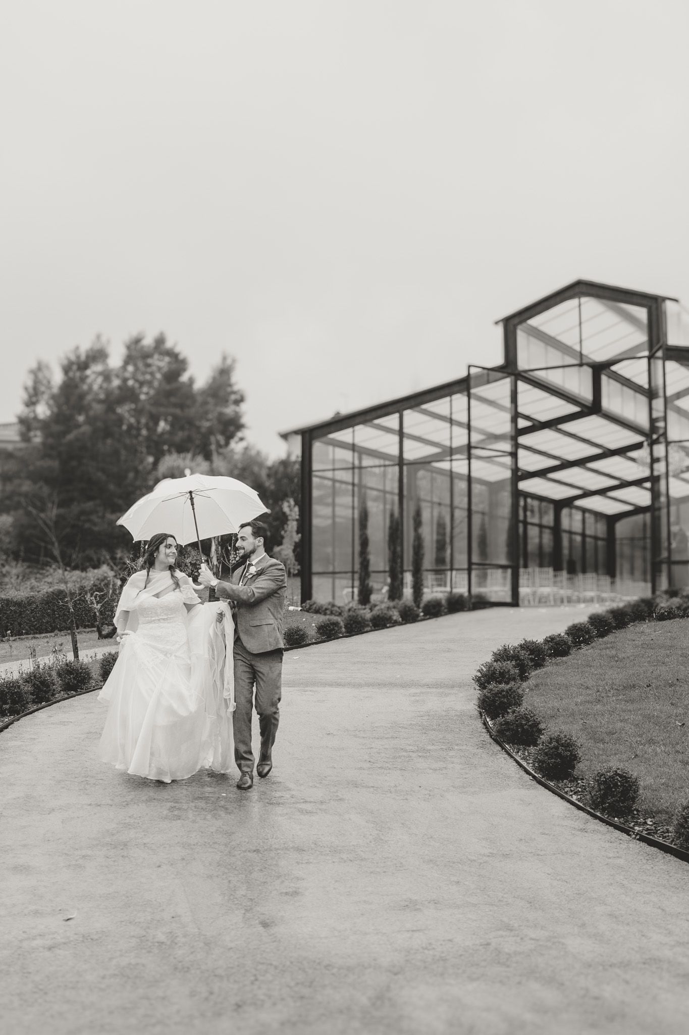Couple strolls along pathway beside modern glass greenhouse structure.