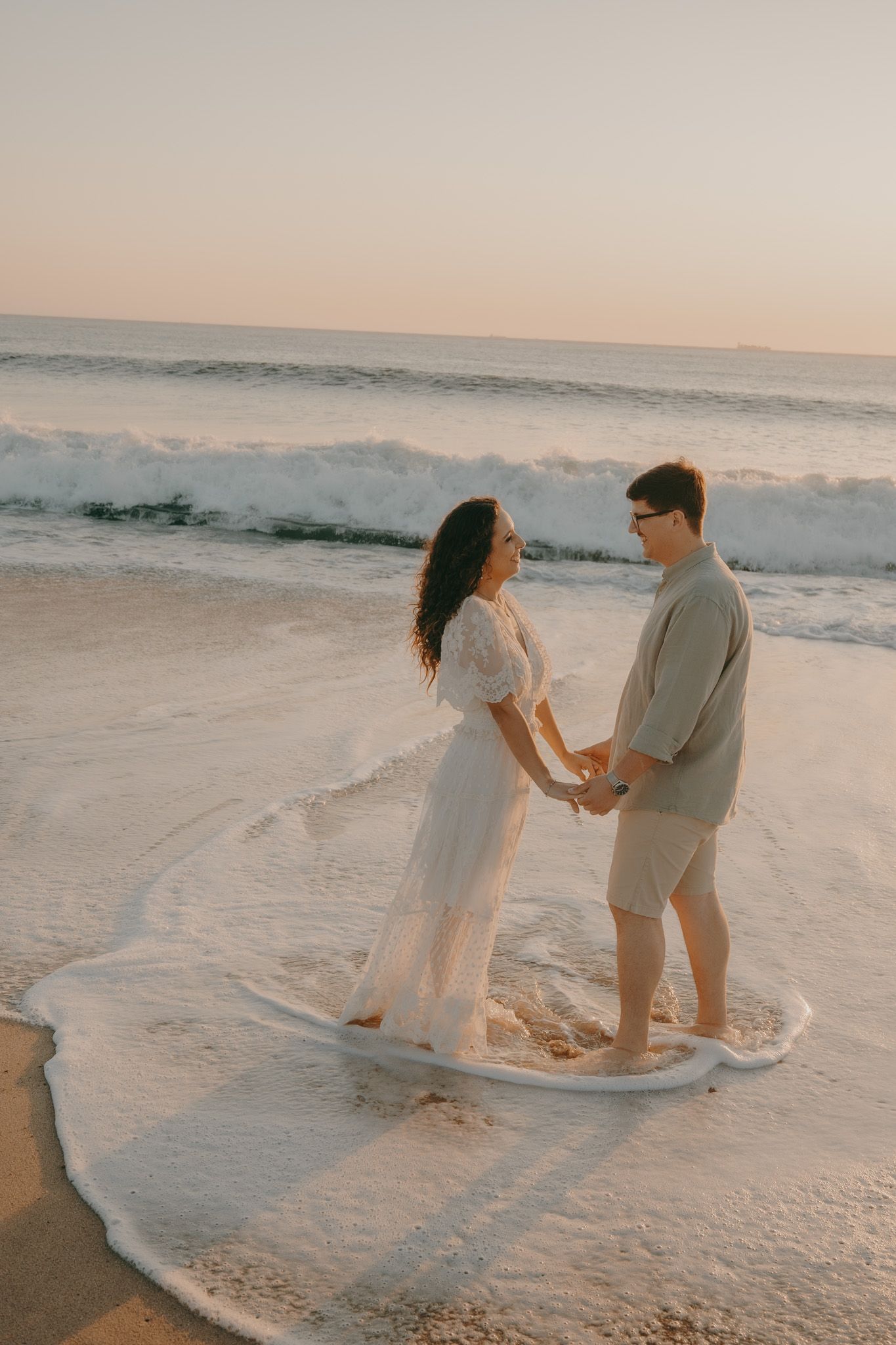 A couple in white and beige attire poses romantically by ocean waves at sunset.