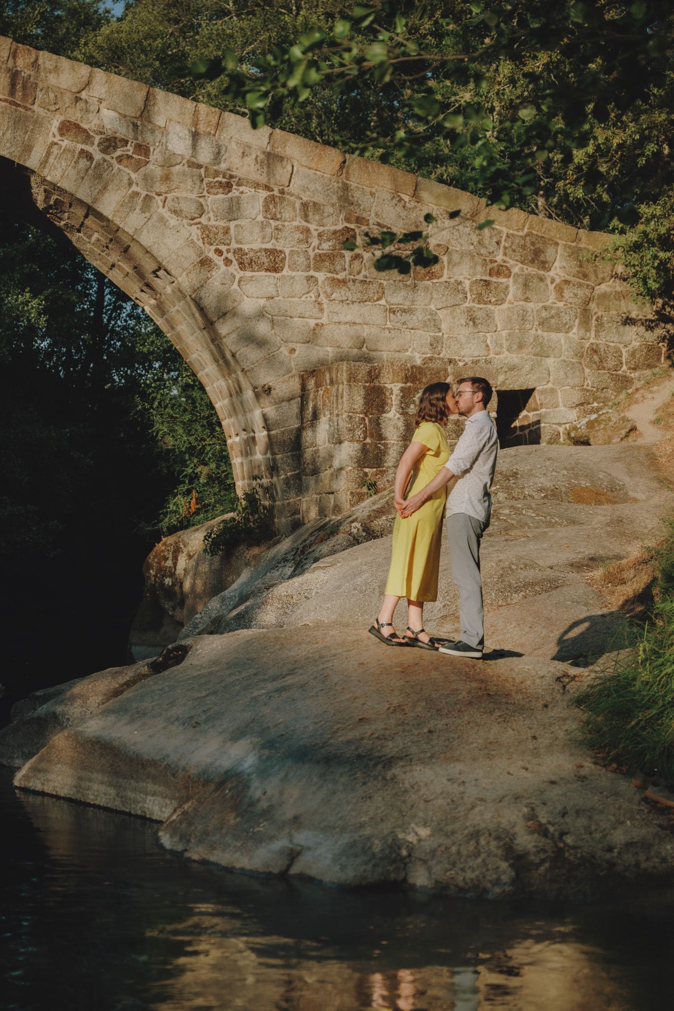 A stone bridge overlooks a peaceful river as sunlight streams through the trees.