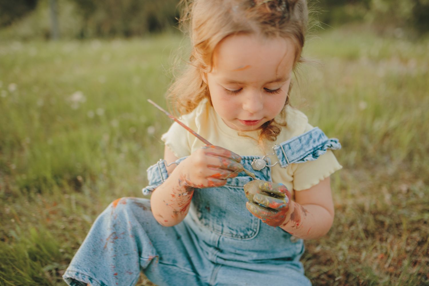 Child in blue overalls reading outdoors among wildflowers and grass.