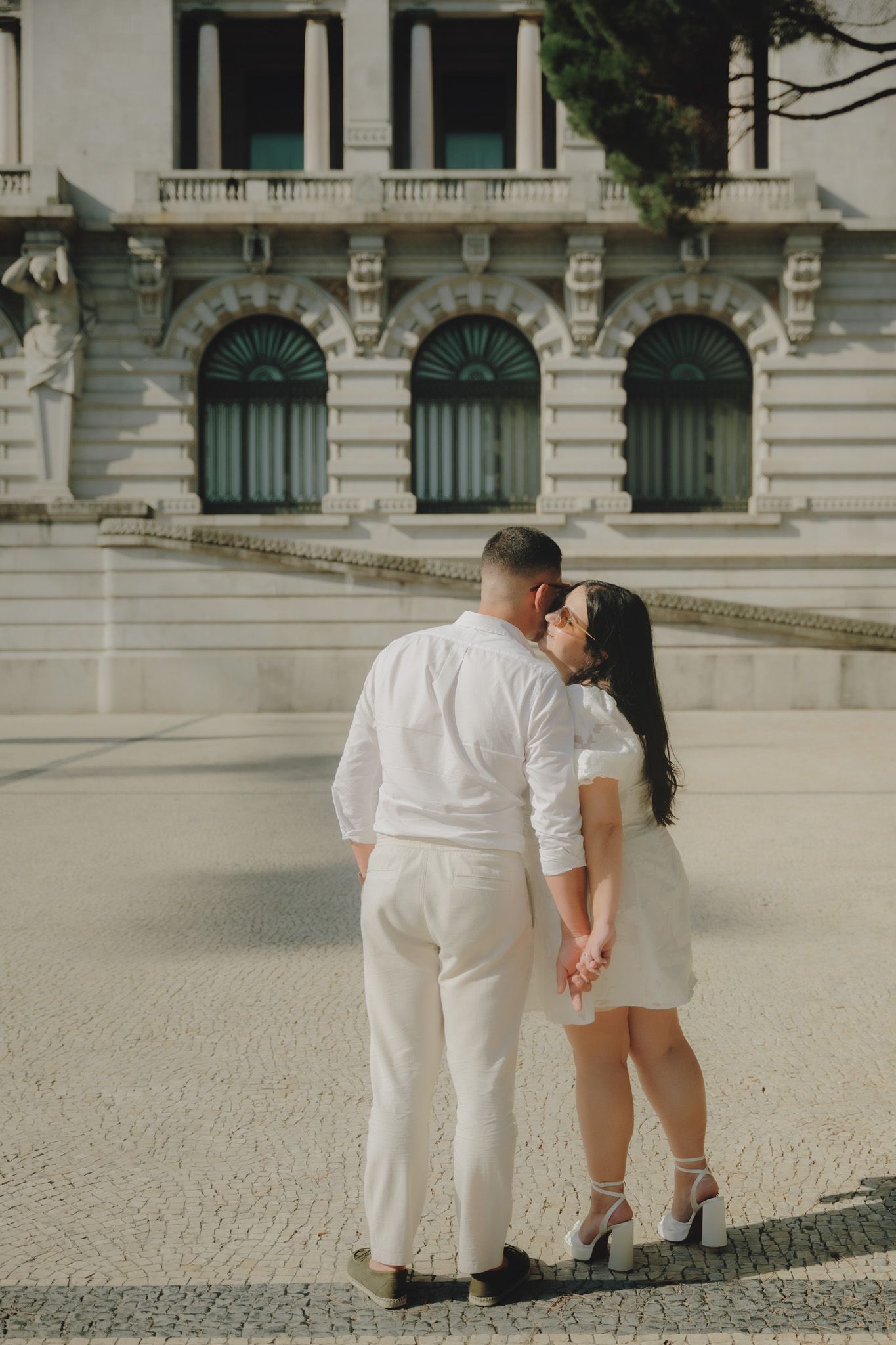 Couple in matching white outfits sharing intimate moment against classical architecture.