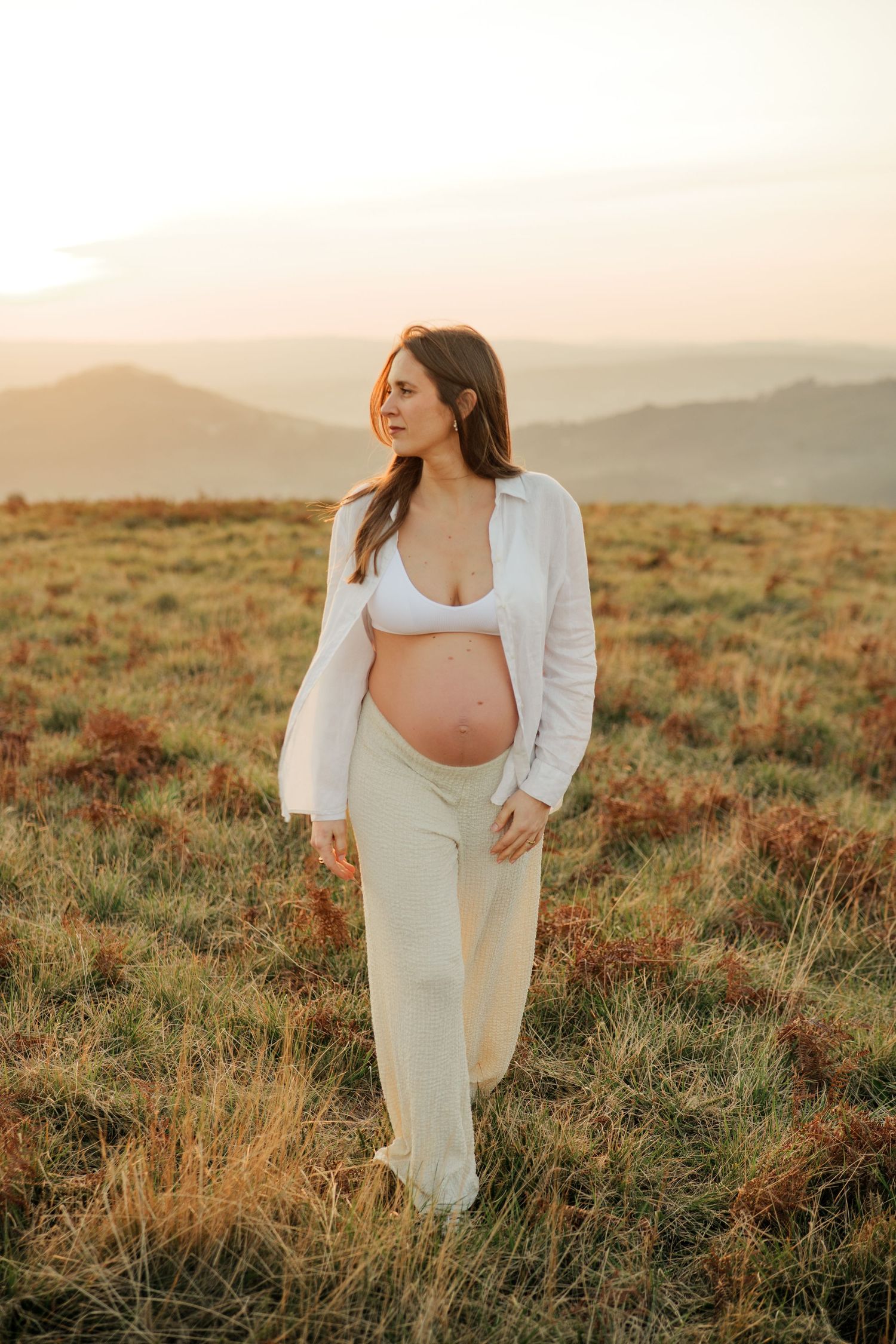 A pregnant woman in white dress poses in a desert landscape at golden hour with mountains in background.
