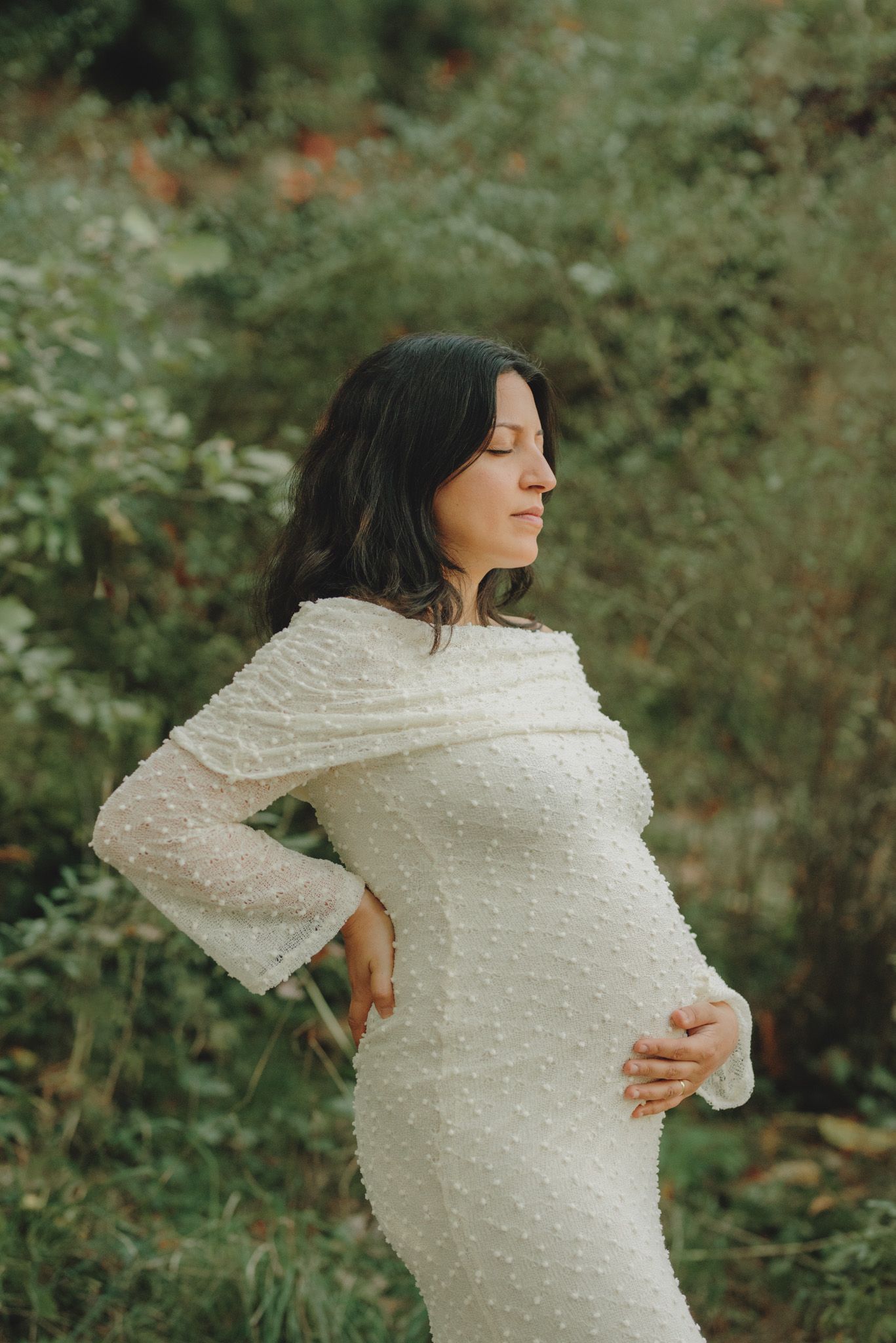 A woman in a textured white dress poses against a backdrop of lush greenery.