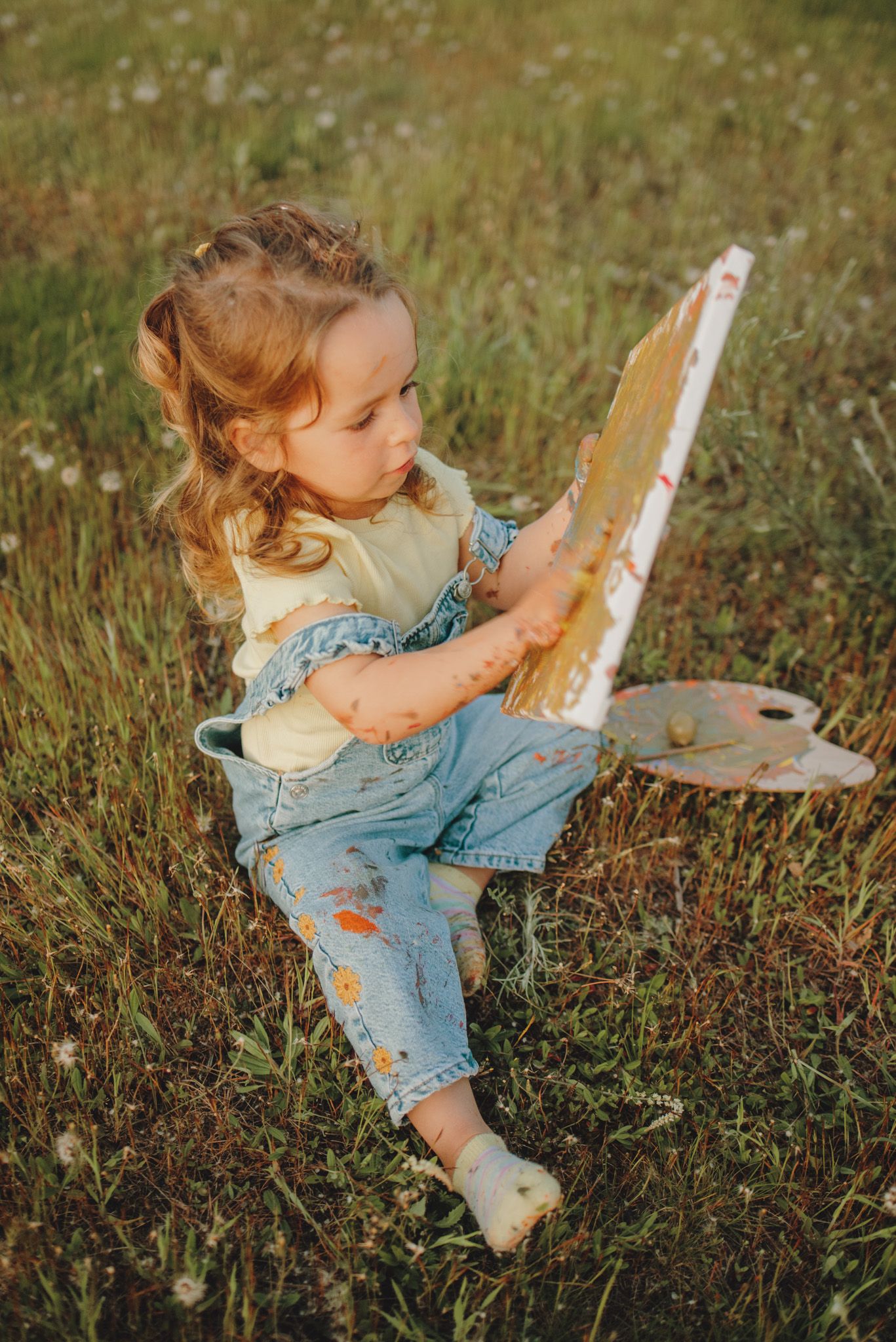 A child in denim overalls lays in a grassy field while reading a book in warm sunlight.