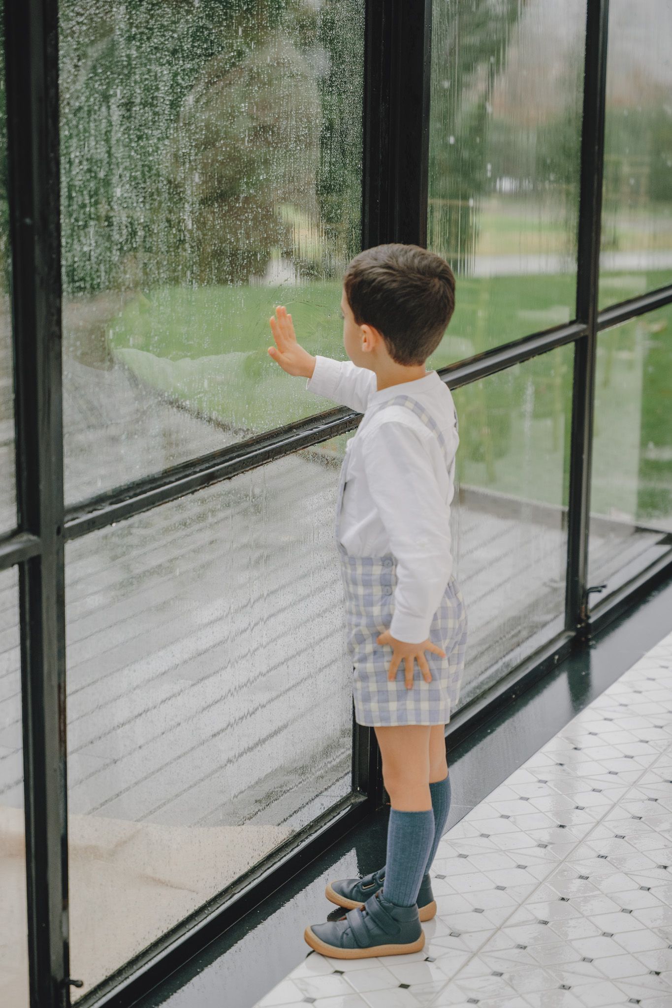 Small child in white shirt and plaid shorts stands by large window looking outside on rainy day.