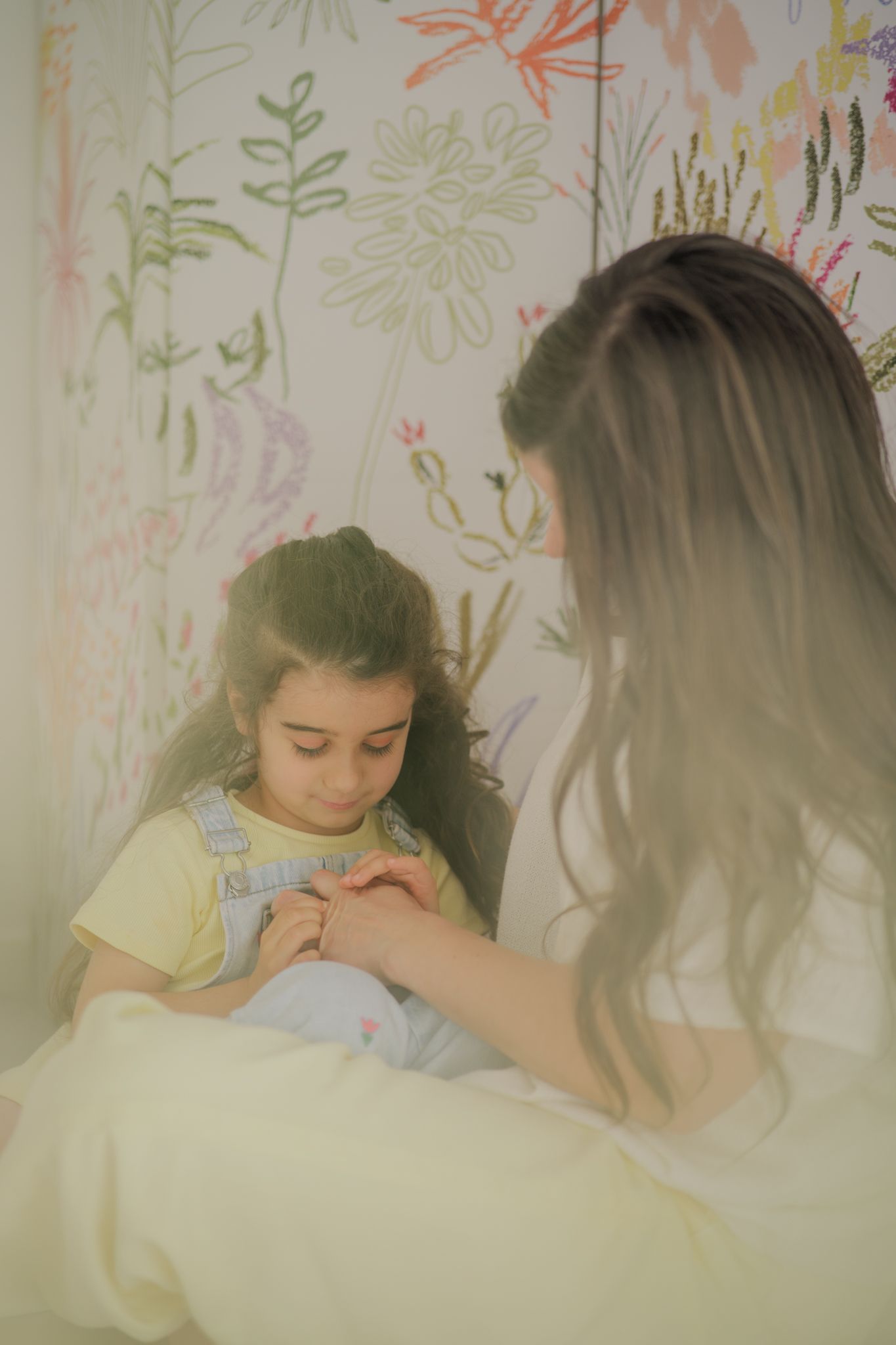A woman tenderly holds a young girl's hands in a floral wallpaper room, both wearing casual clothes.