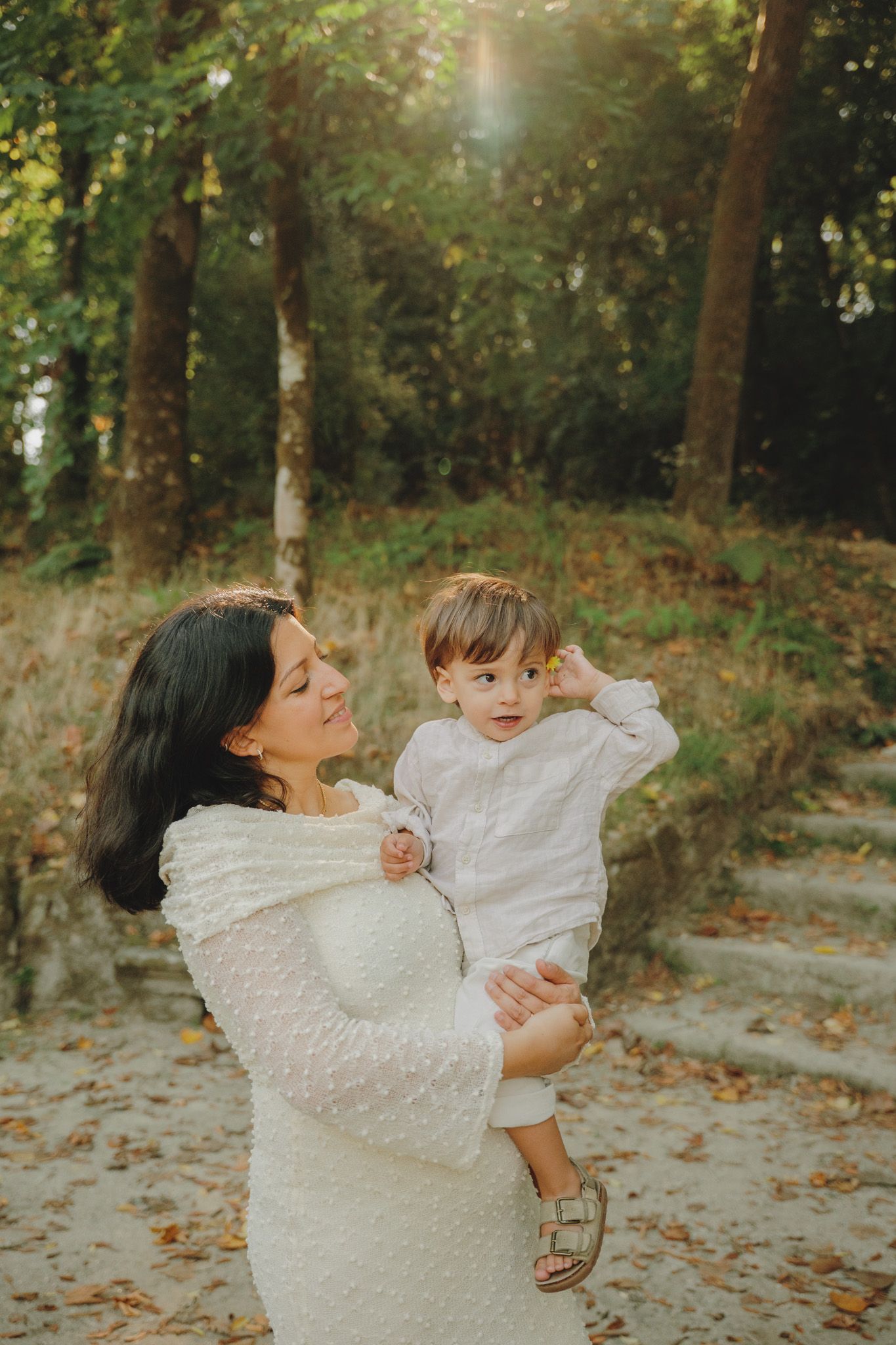 Mother and toddler in matching white outfits share a tender moment in autumn forest setting.