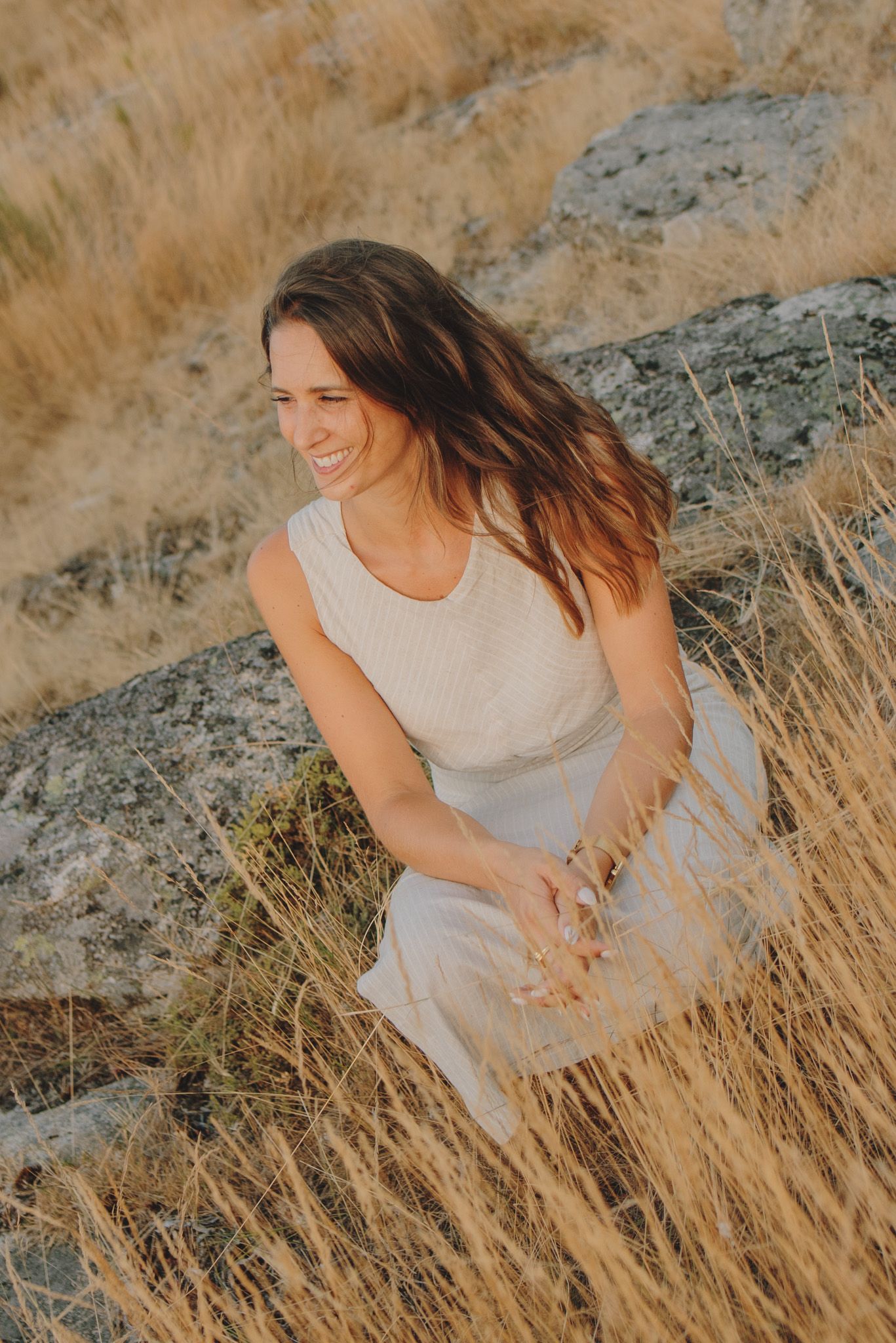 Woman sitting in tall golden grass during sunset with mountains in the distance.