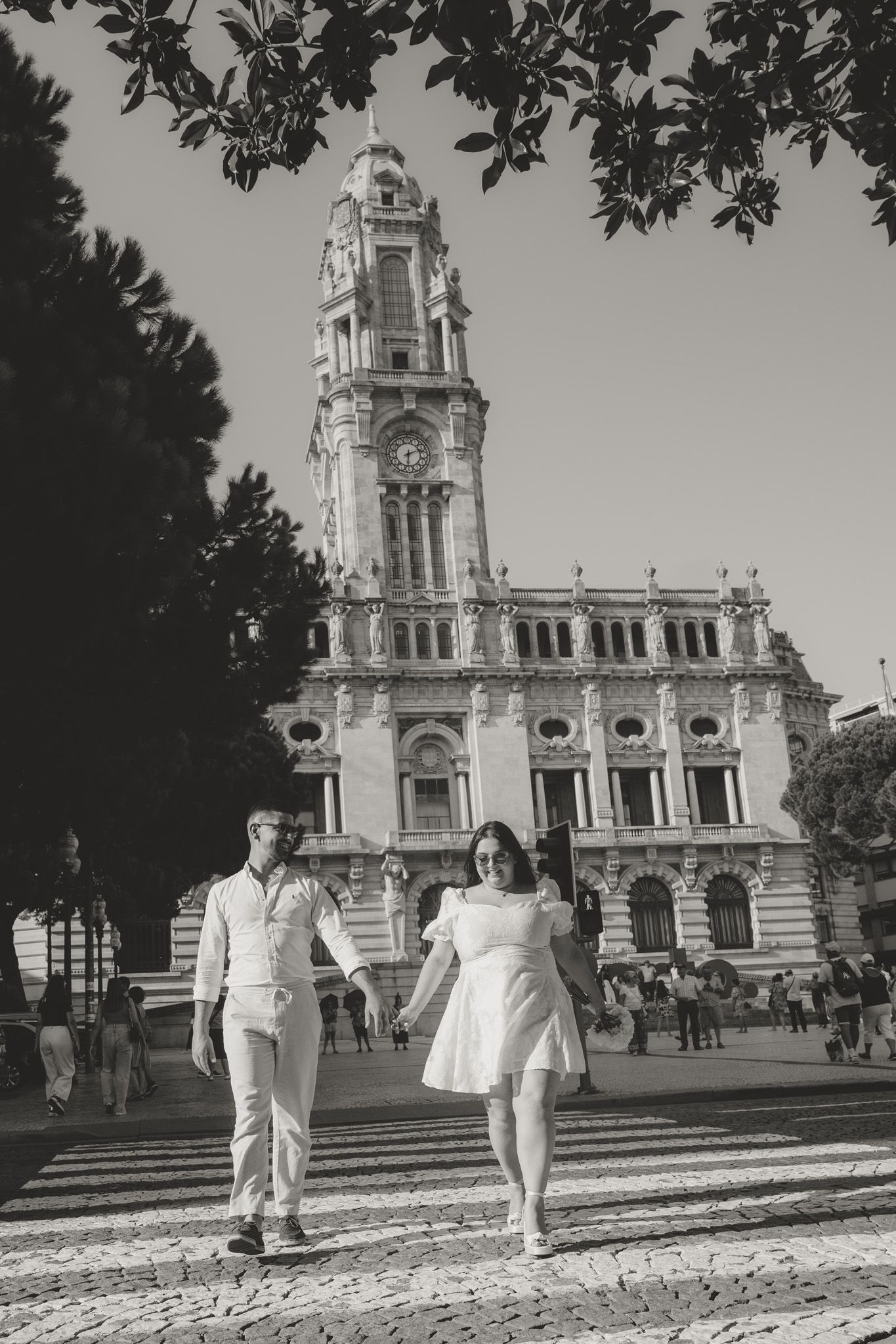 A couple strolling in front of an ornate historic building with a clock tower on a sunny day.
