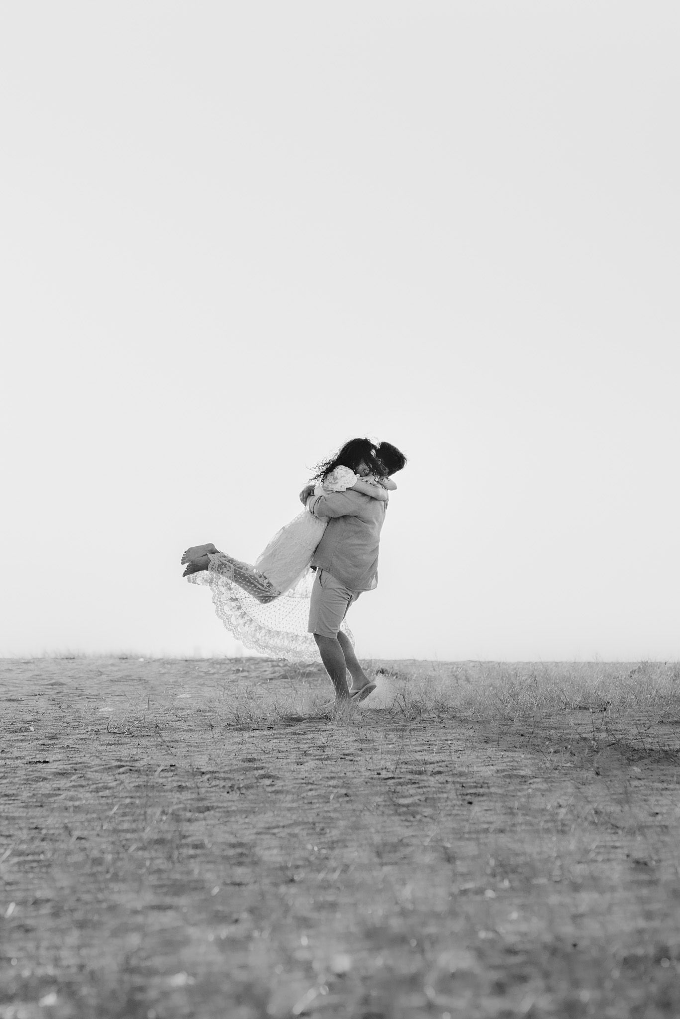 Black and white photo of a couple playfully dancing on a hillside.