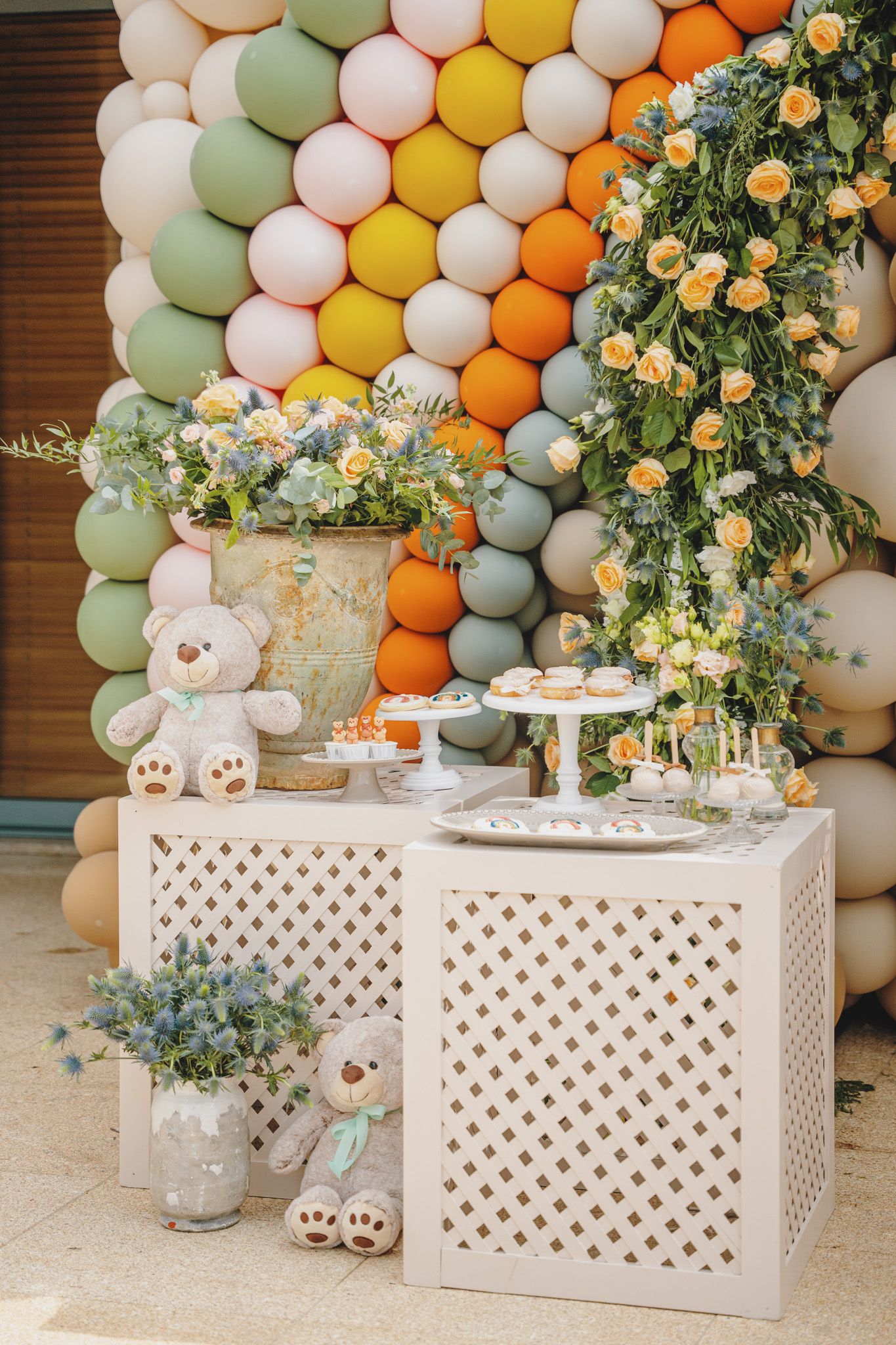 White lattice display tables with floral arrangements and teddy bears against a colorful balloon backdrop.