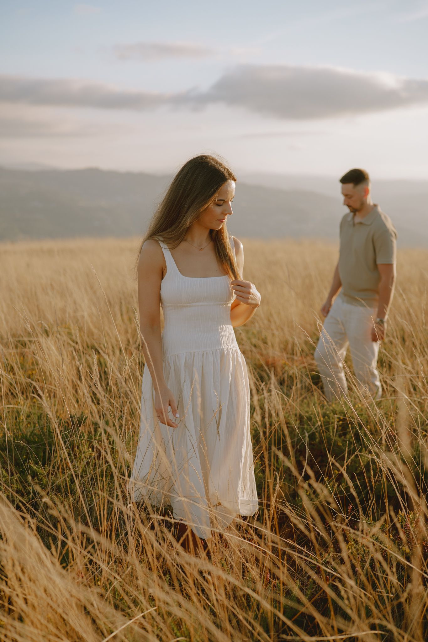 Couple in white and grey clothing walking through tall golden grass at sunset.