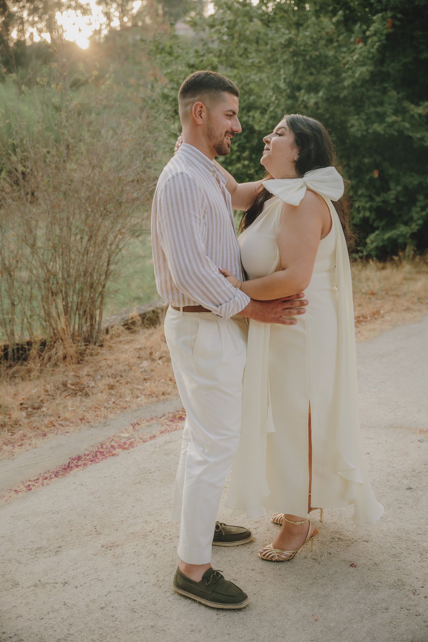 A romantic outdoor photoshoot featuring a couple in white outfits posing on a dirt path at sunset.