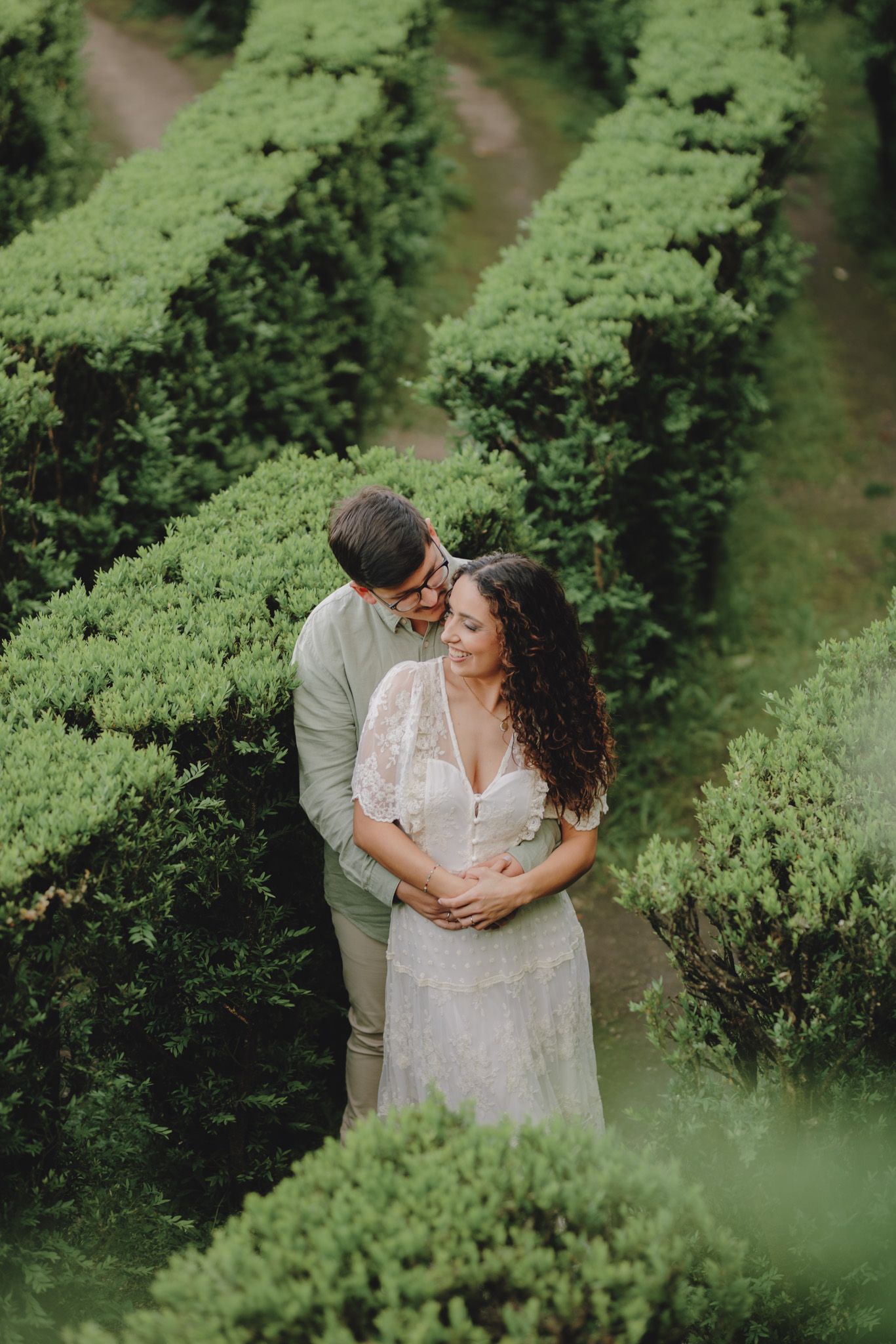 A couple shares an intimate moment among lush green hedgerows in a garden setting.