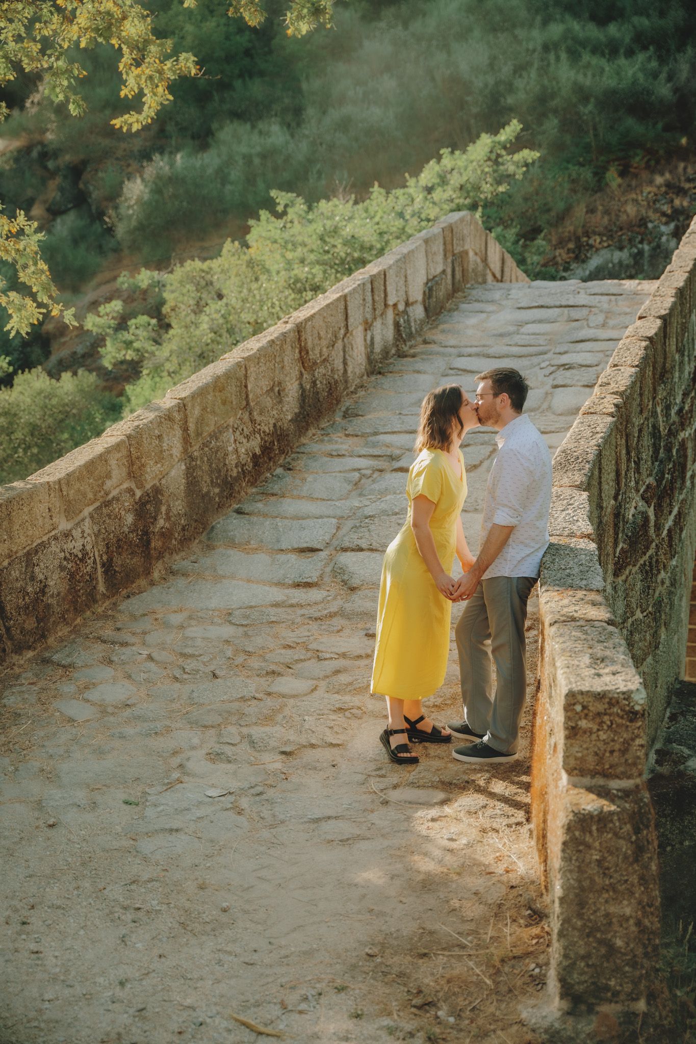 A couple shares a romantic moment on a stone bridge path surrounded by greenery.
