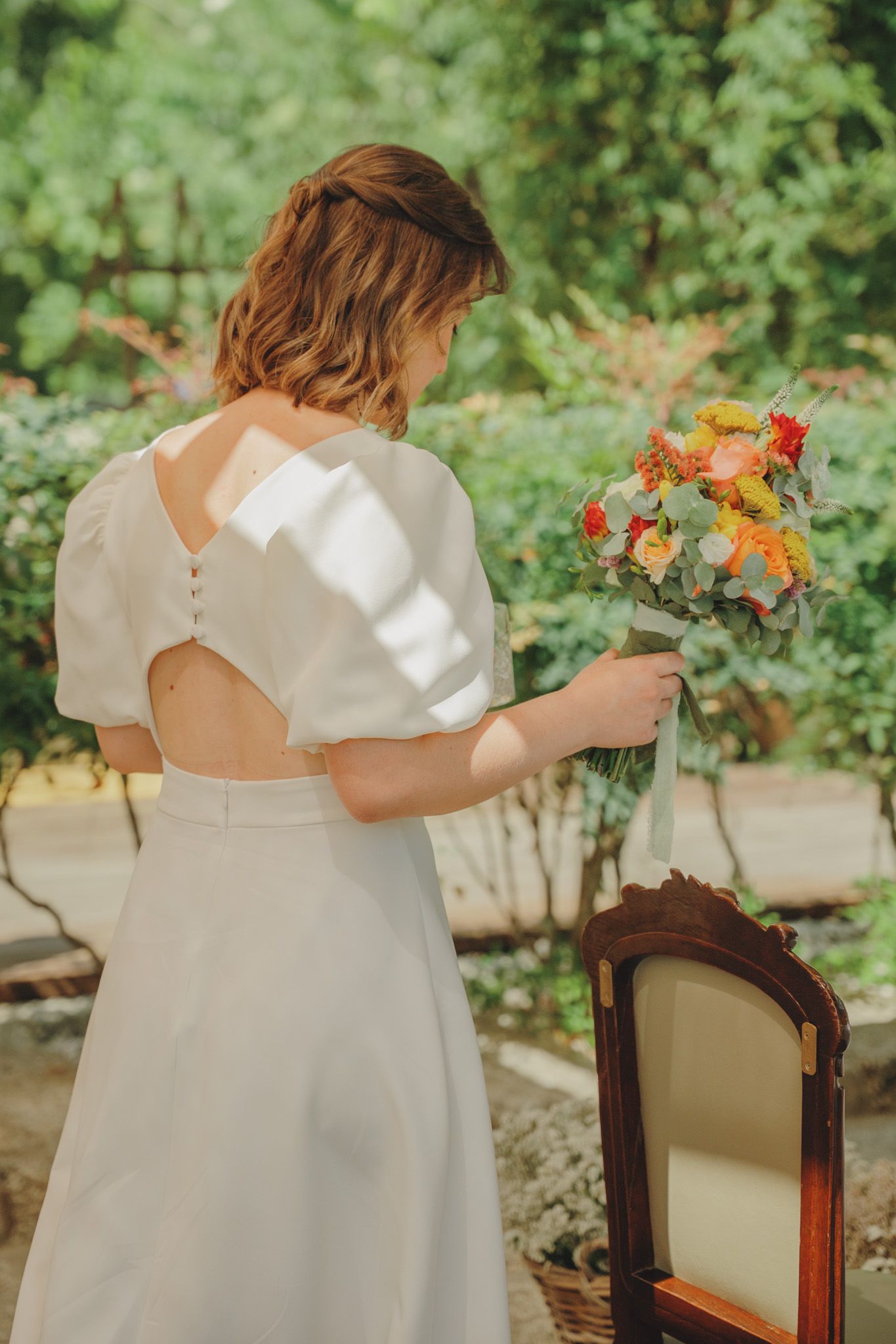 Back view of a white wedding dress with delicate details and floral bouquet against garden backdrop.