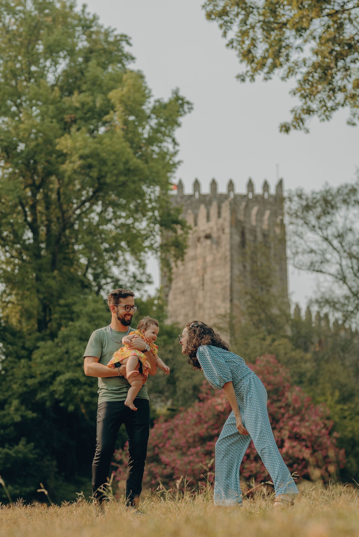A couple plays with their small dog in a grassy field with a stone church tower visible in the background.