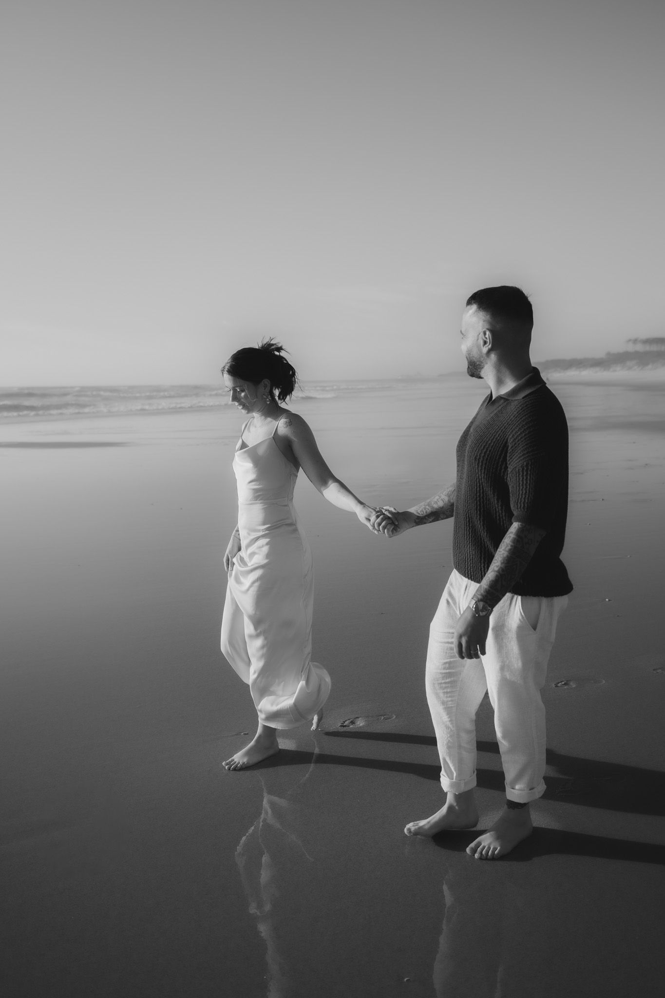 Dramatic black and white photo of two people holding hands on a serene beach at dusk.